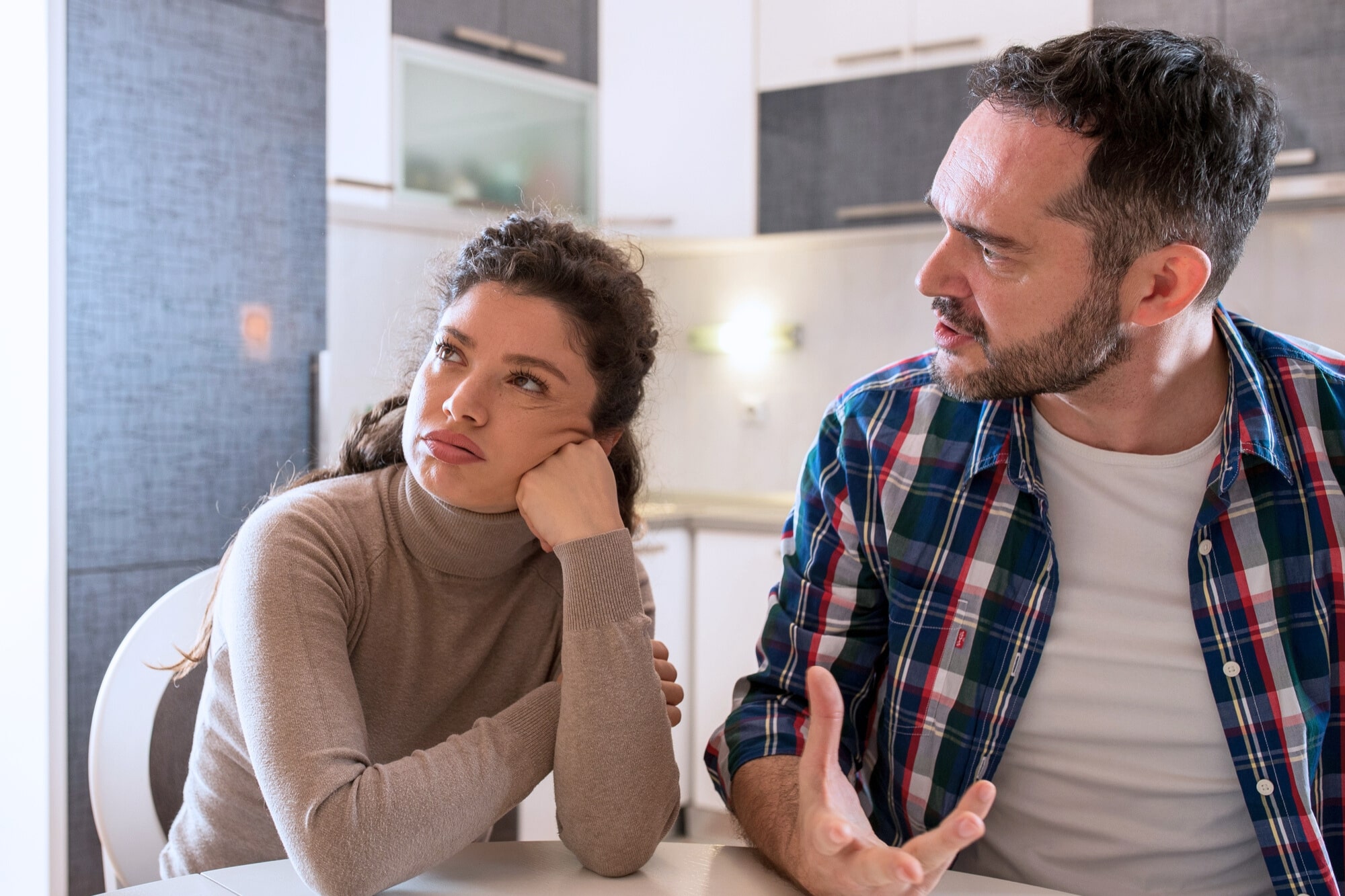 A woman sits at a table looking away with a bored or frustrated expression, resting her chin on her hand, while a man next to her gestures and speaks, appearing upset. They are in a modern kitchen.