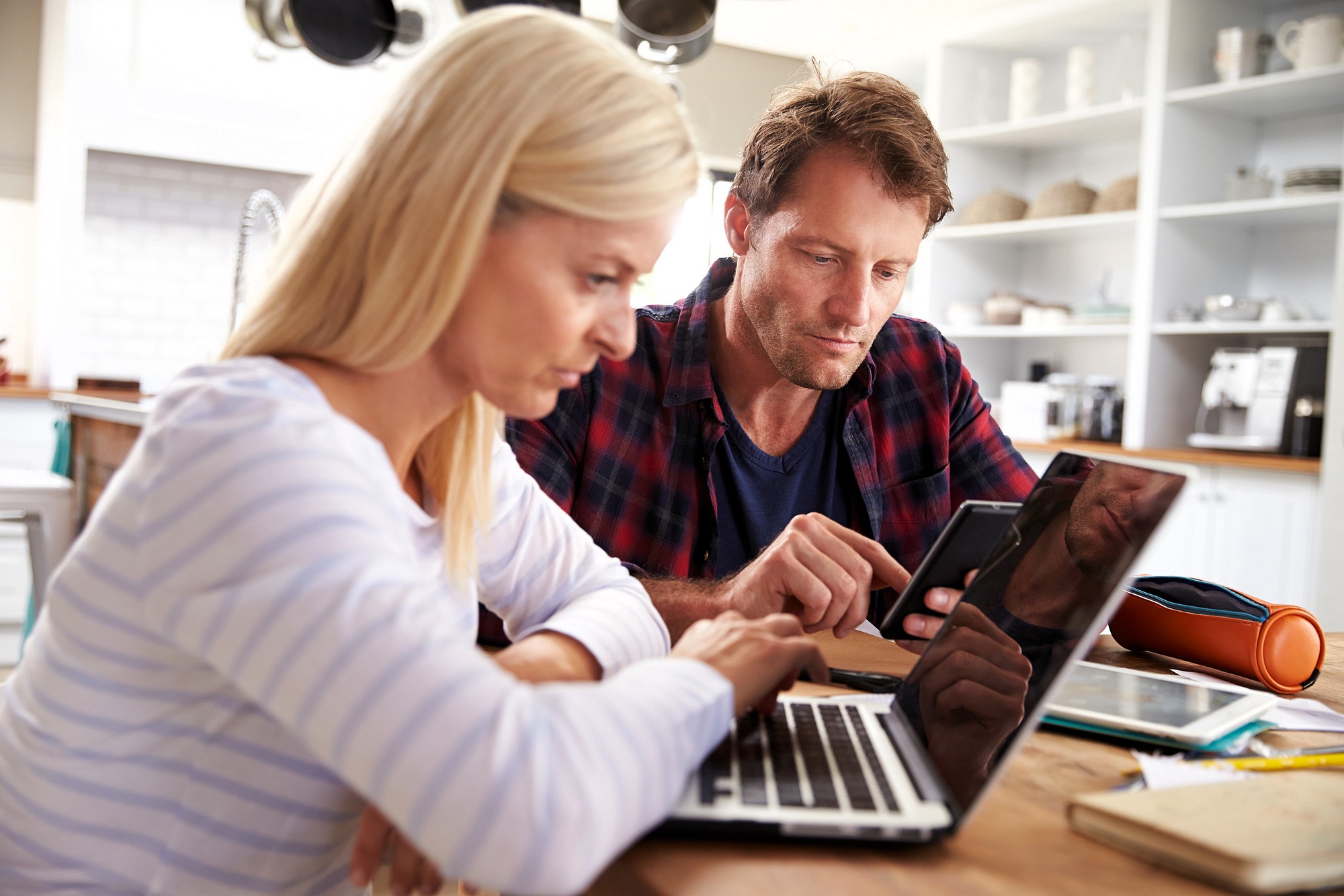 A woman and a man sit at a kitchen table, looking at a laptop and a smartphone. The woman types on the laptop while the man checks his phone, both appearing focused and engaged in their work.