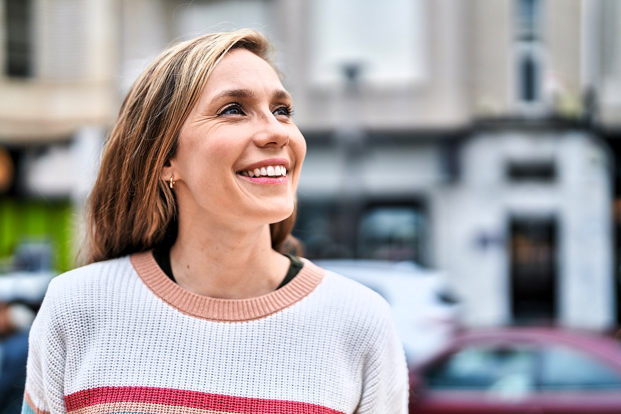 A woman with long hair smiles and looks up while standing outdoors. She is wearing a white sweater with pink and red stripes, and the background shows a blurred city street scene.