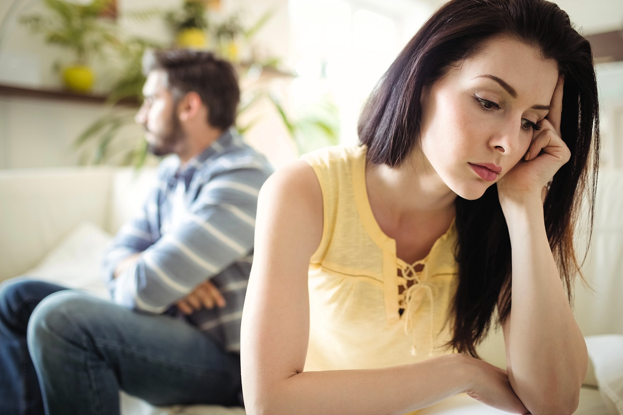 A woman sits in the foreground looking upset with her head resting on her hand, while a man sits in the background with his arms crossed, both appearing to be in the middle of an argument or disagreement.