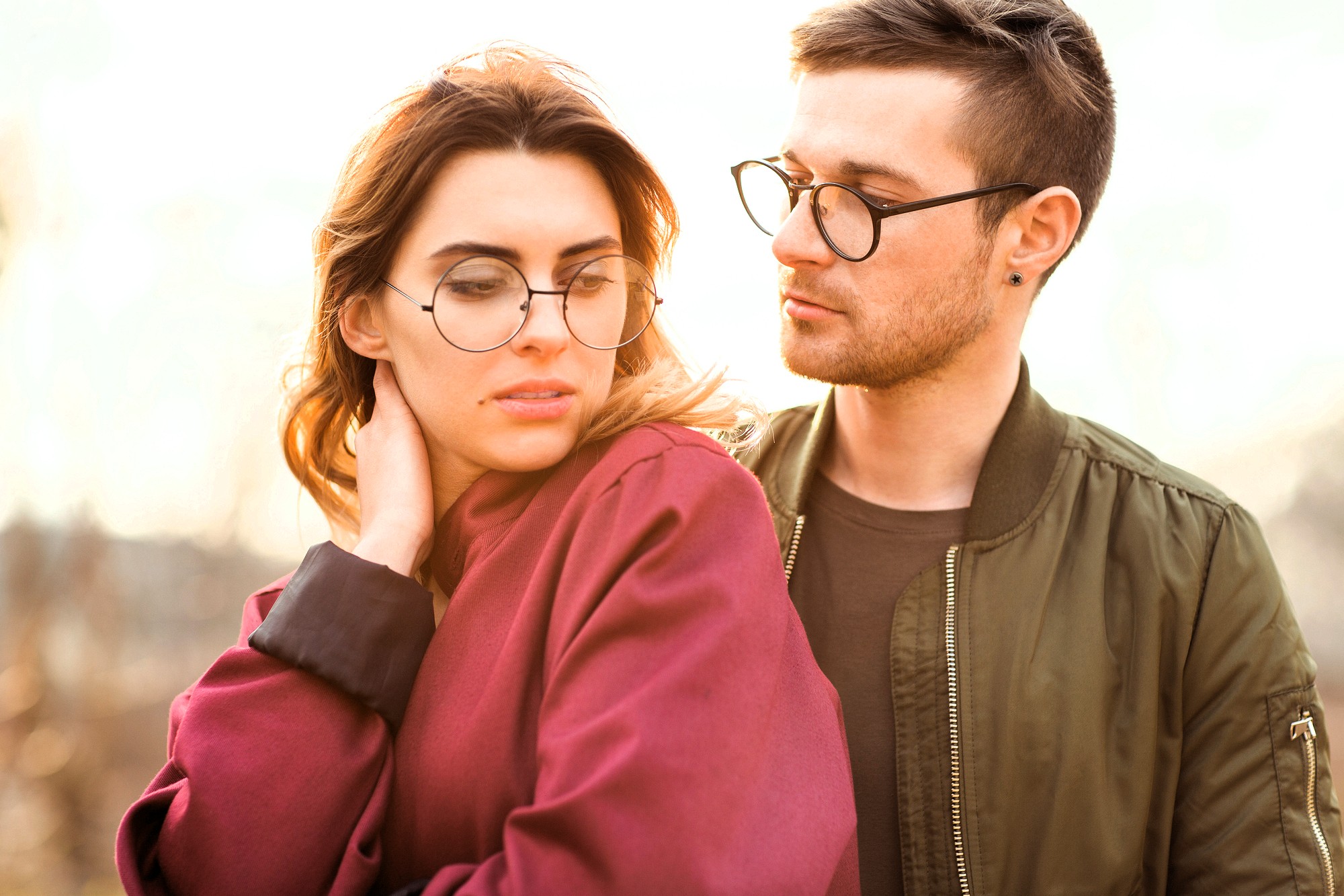 A young woman in a red jacket and glasses looks down while a young man in a green jacket and glasses stands close beside her, gazing at her with a soft expression. They are outdoors in natural light.