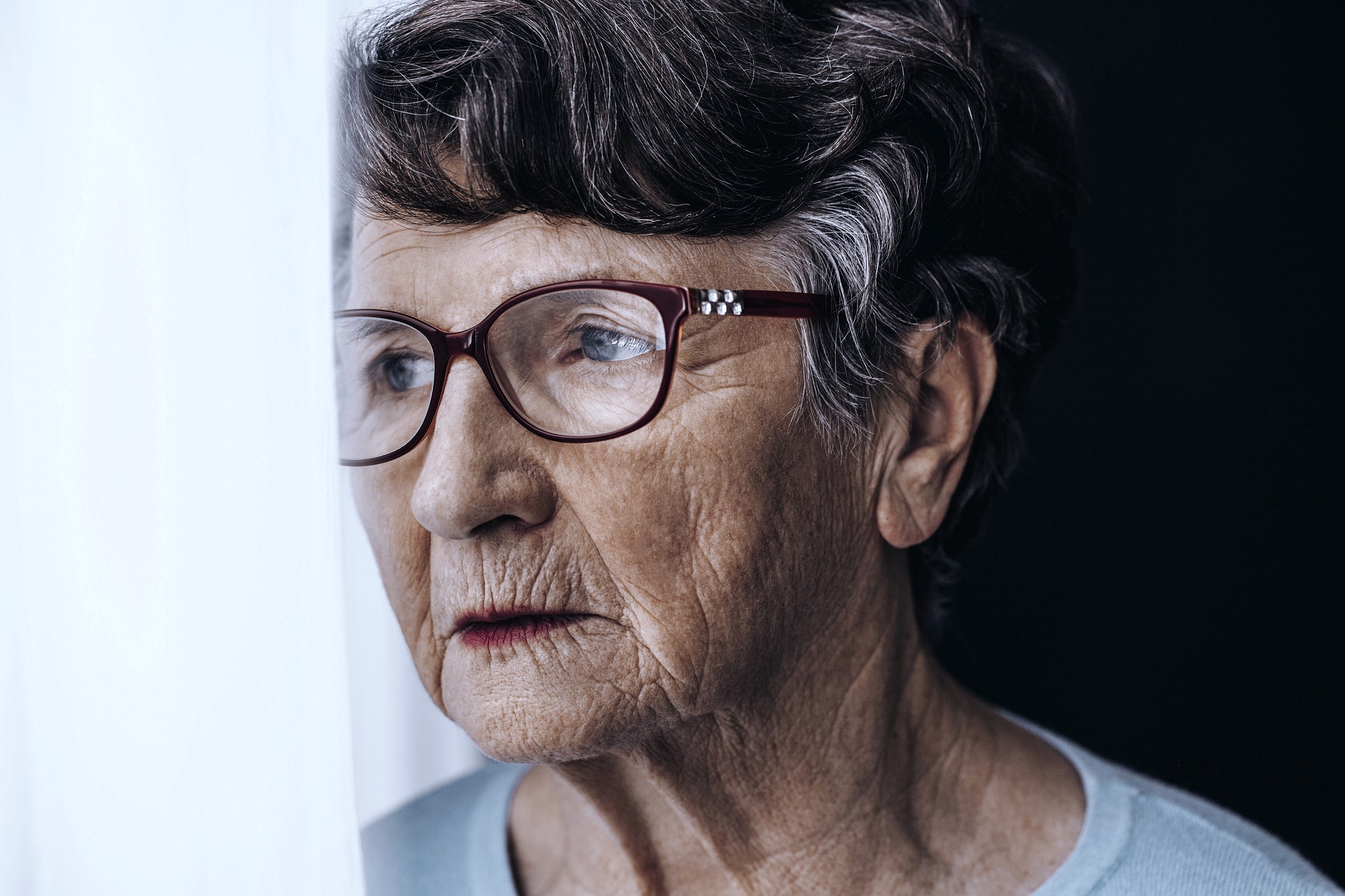 An elderly woman with short gray hair and glasses looks thoughtfully out a window, her face partially illuminated by soft light, with a neutral expression.