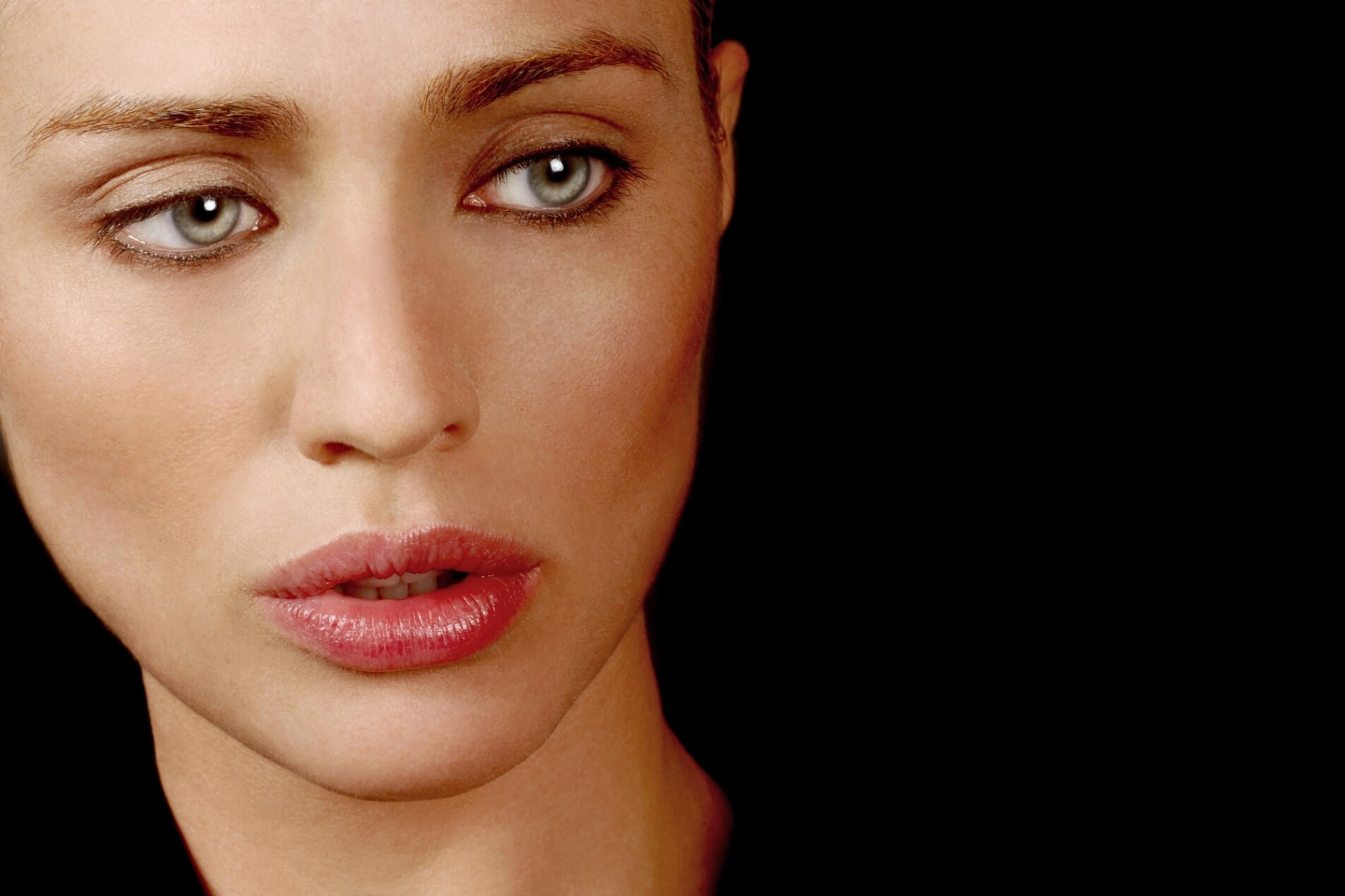A close-up of a woman with pale skin, blue eyes, and pink lips looking slightly to the side against a black background.