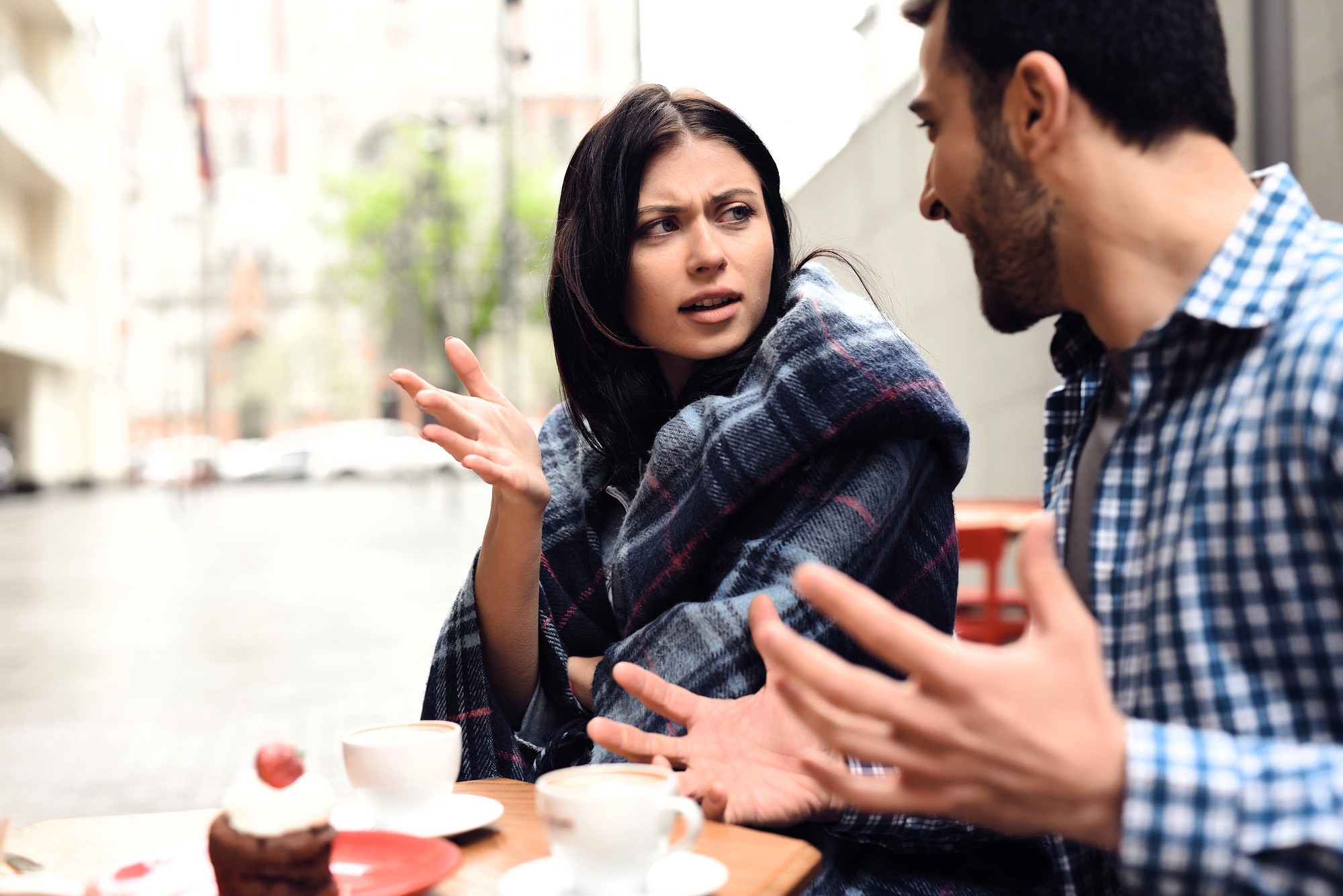 A woman wrapped in a blanket looks frustrated while talking to a man at an outdoor café table with coffee and dessert. Both have expressive hand gestures, suggesting a serious conversation.