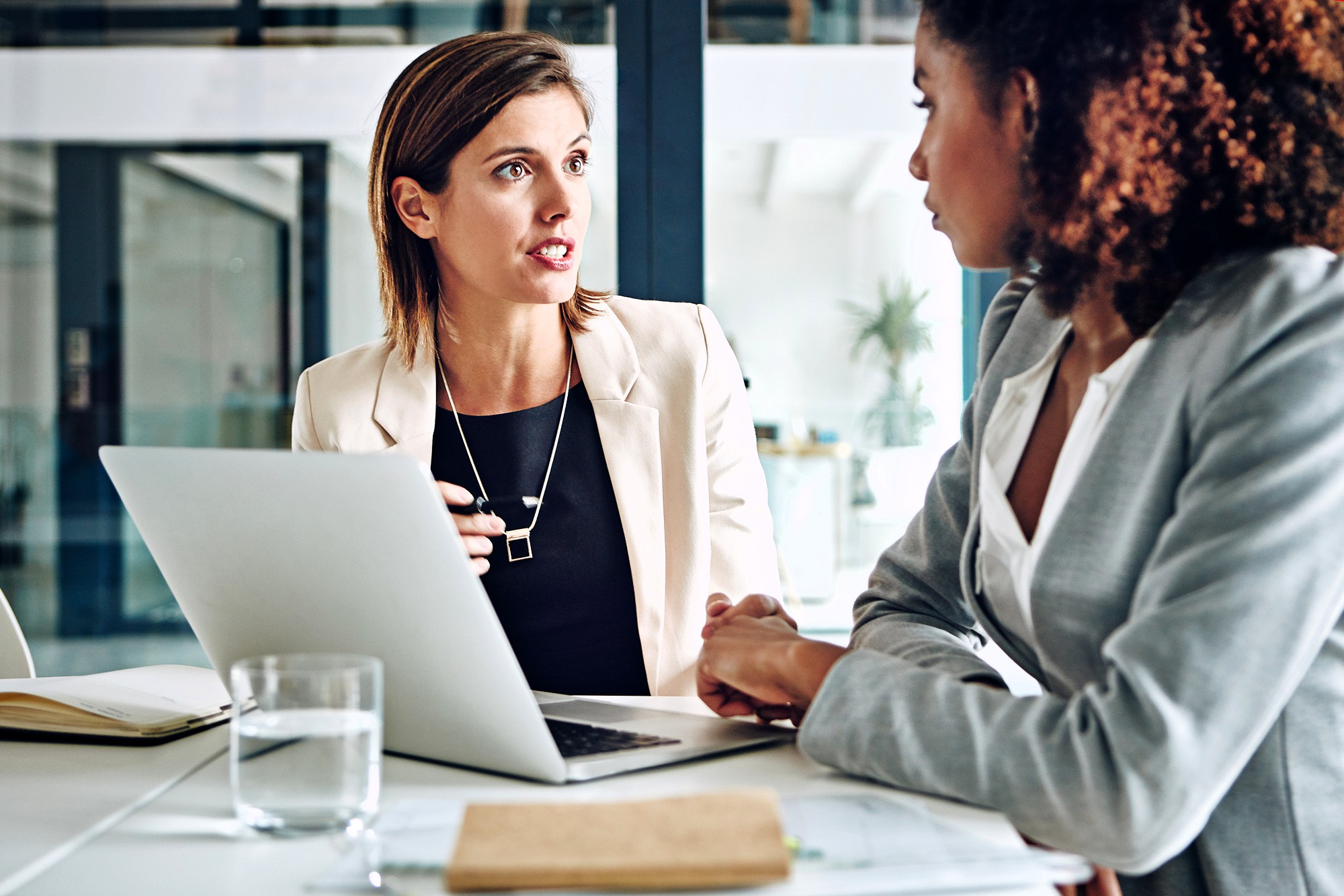 Two women in business attire are sitting at a desk with a laptop and documents, engaged in a serious conversation in a modern office setting.