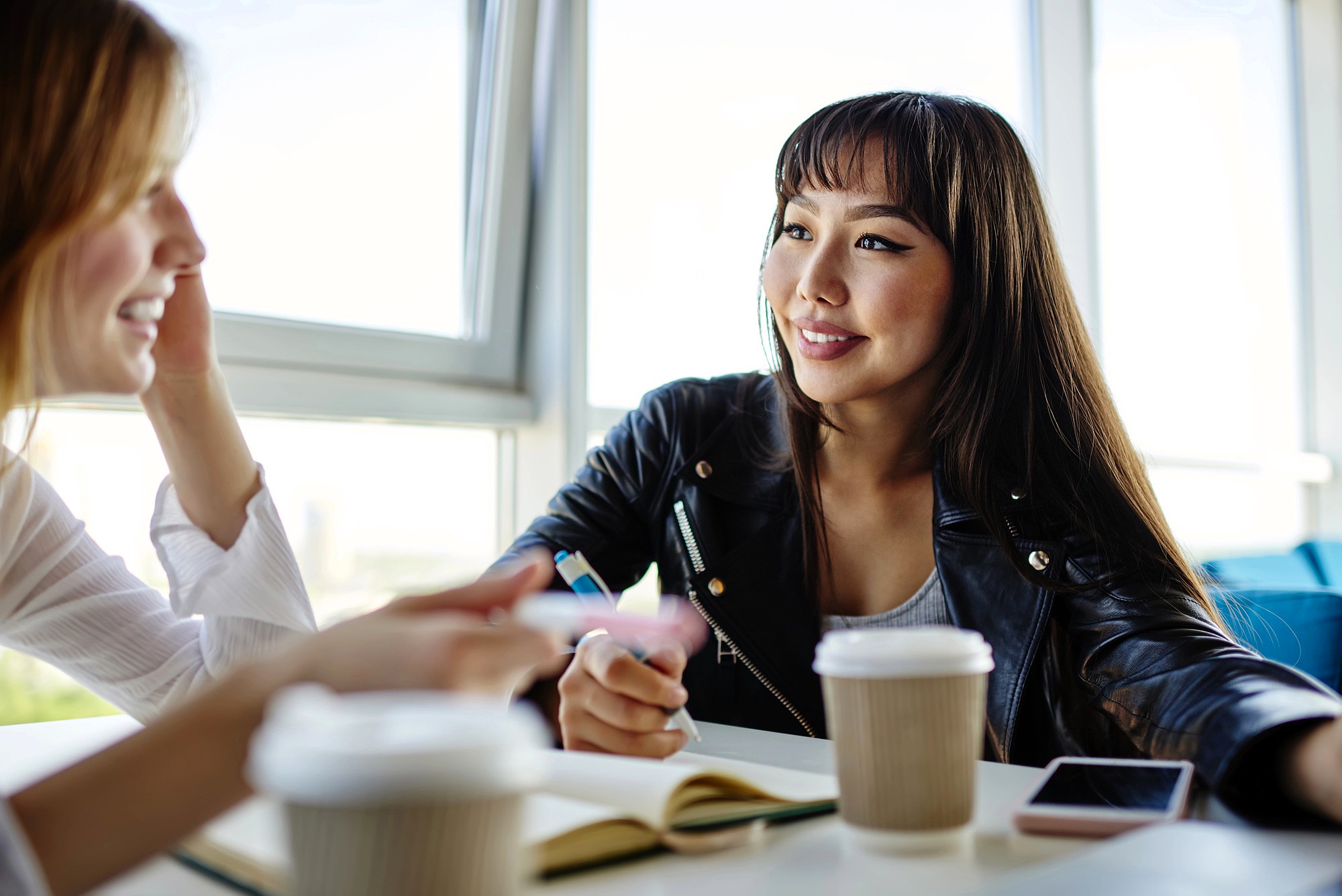 Two women sit at a table in a bright cafe, talking and smiling. They have coffee cups, a notebook, and a smartphone on the table. One woman wears a black jacket; the other gestures with her hand while speaking.