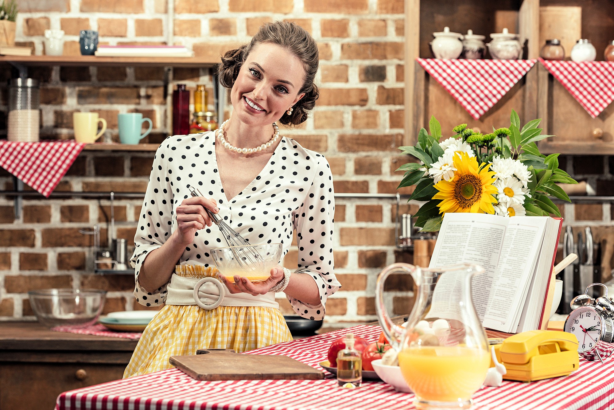 A woman in retro clothing whisks eggs in a kitchen decorated with red checkered cloths and flowers. There's an open cookbook, a glass pitcher, and a vintage phone on the counter. She smiles at the camera.