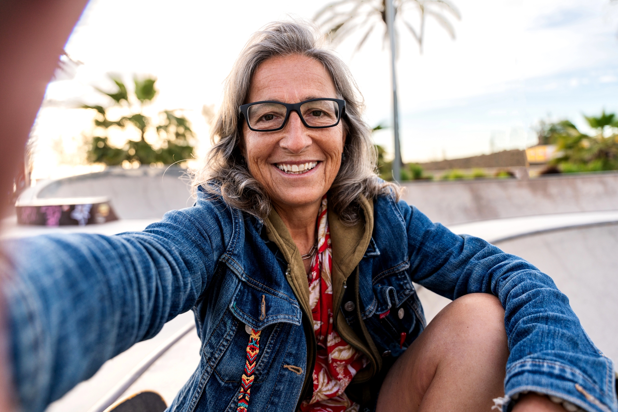Smiling older woman with long gray hair and glasses takes a selfie at a skatepark, wearing a denim jacket and patterned shirt. Palm trees and a bright sky are visible in the background.