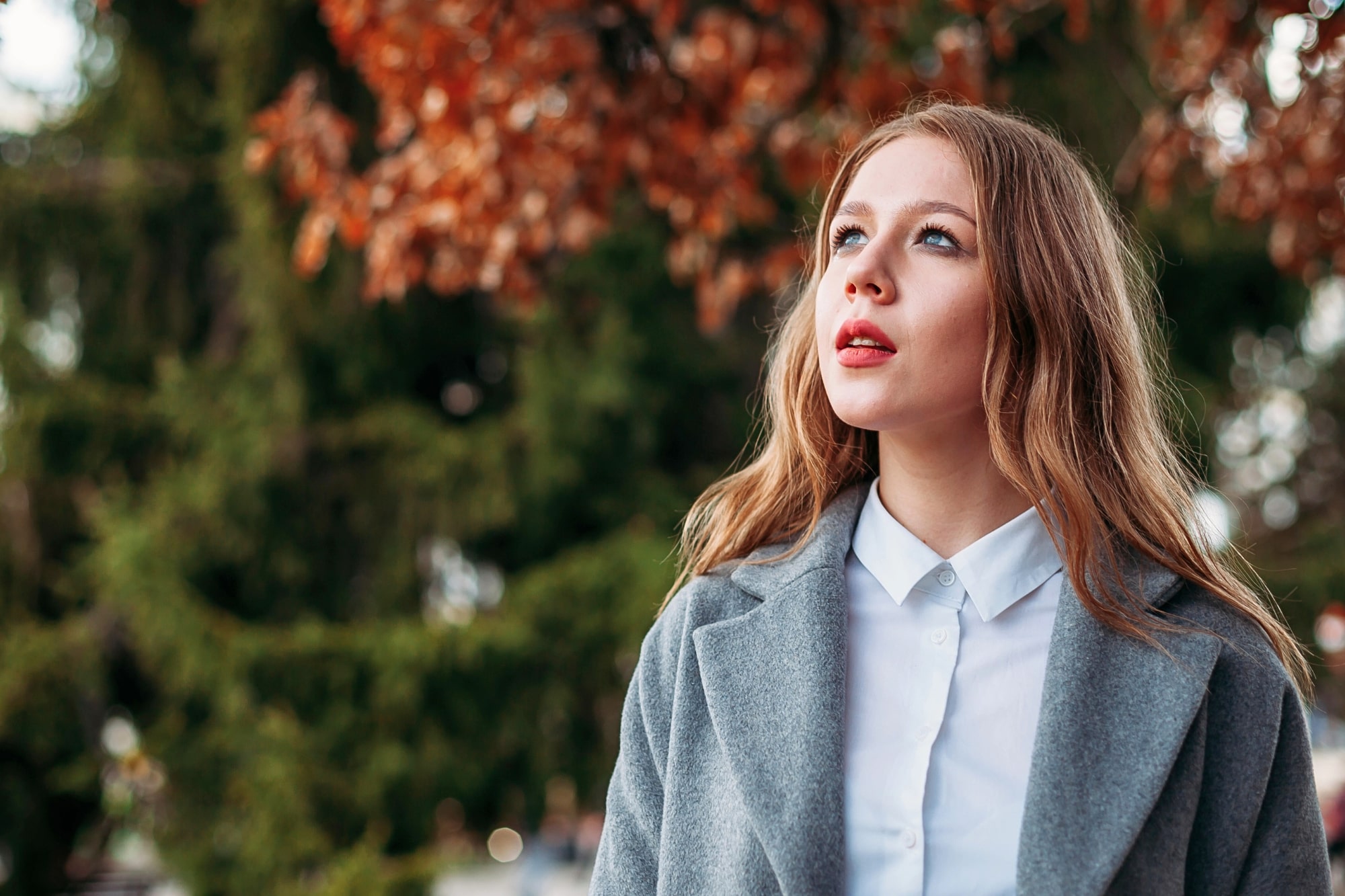 A young woman with long, light brown hair wearing a gray coat and white shirt looks up thoughtfully outdoors. Autumn leaves and green trees are blurred in the background.