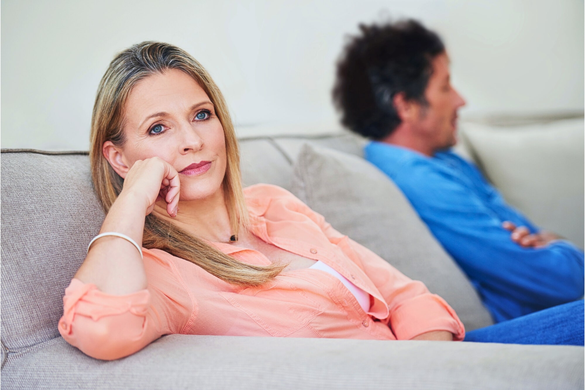 A woman in a peach shirt sits on a couch, looking thoughtful and slightly concerned, with her hand resting on her chin. In the background, a man in a blue shirt sits turned away, appearing distant.