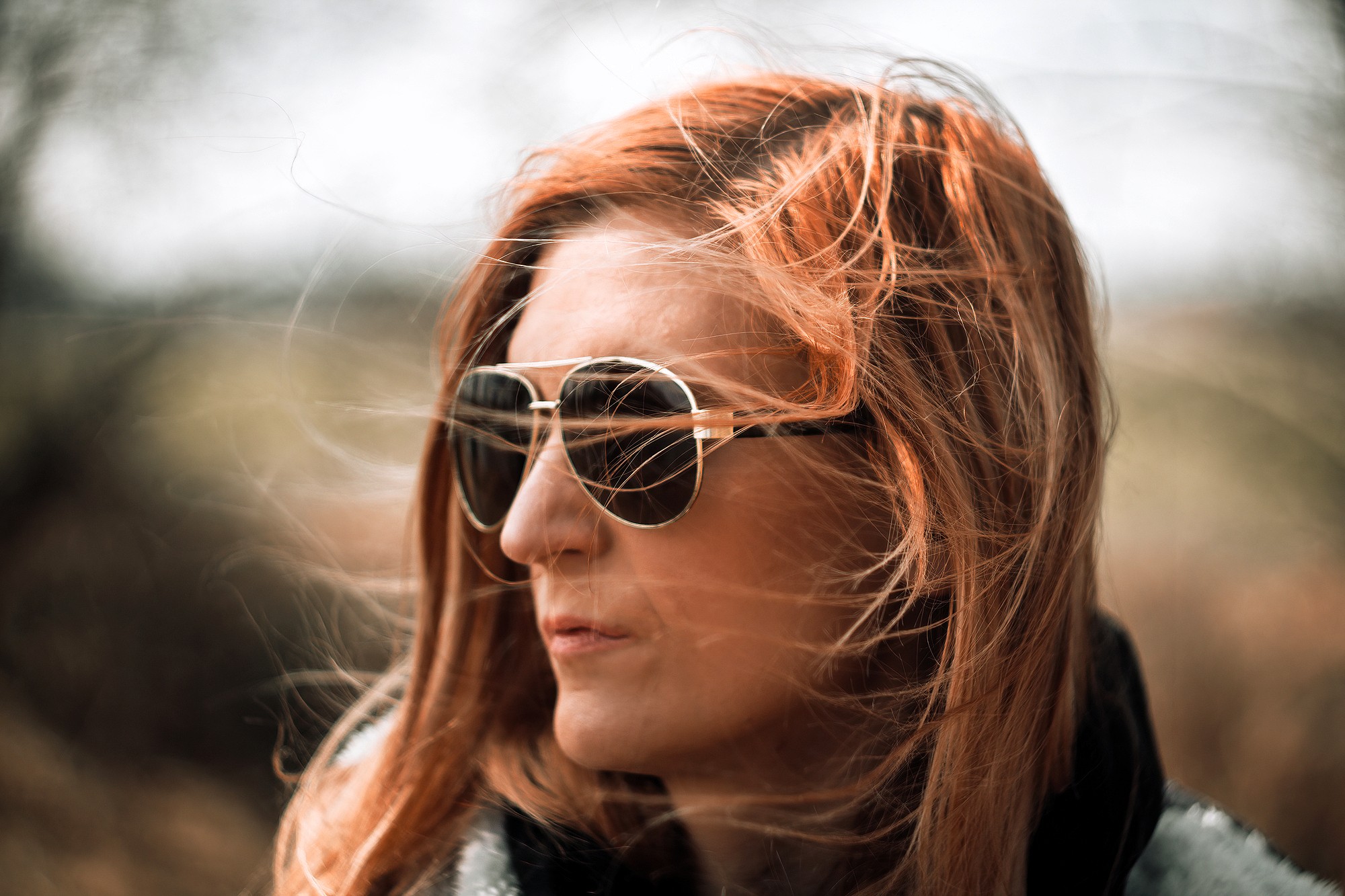 A woman with long, windblown red hair wearing aviator sunglasses looks off to the side outdoors. The background is blurred, highlighting her face and sunglasses.
