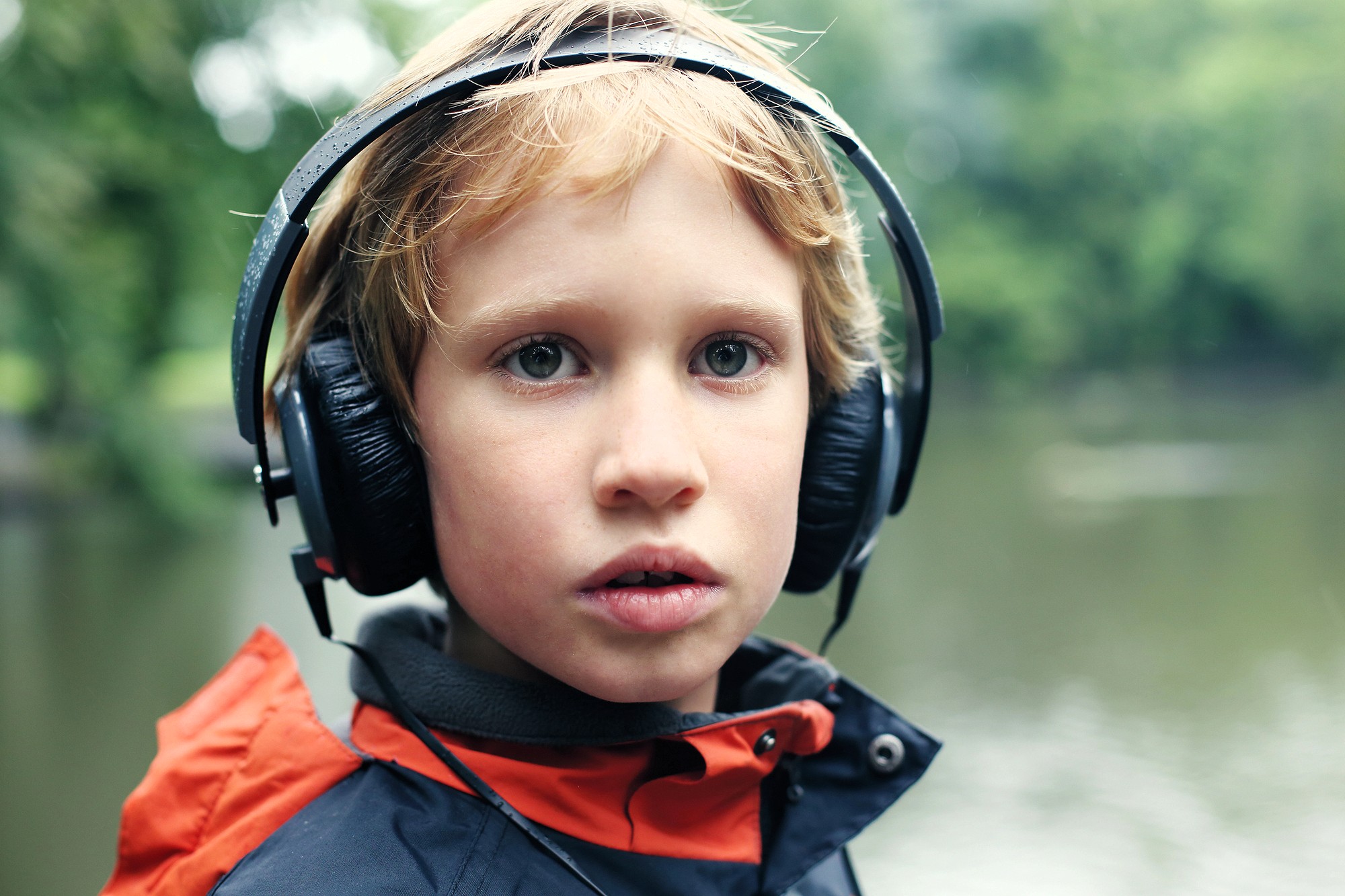 A young child with blonde hair wears large black headphones and a navy blue jacket with an orange hood, standing outdoors near a body of water with blurred green trees in the background.