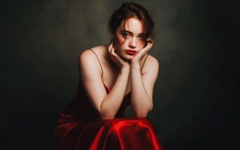 A woman in a red satin dress sits with her elbows on her knees, resting her chin on her hands, looking directly at the camera against a dark, blurred background.