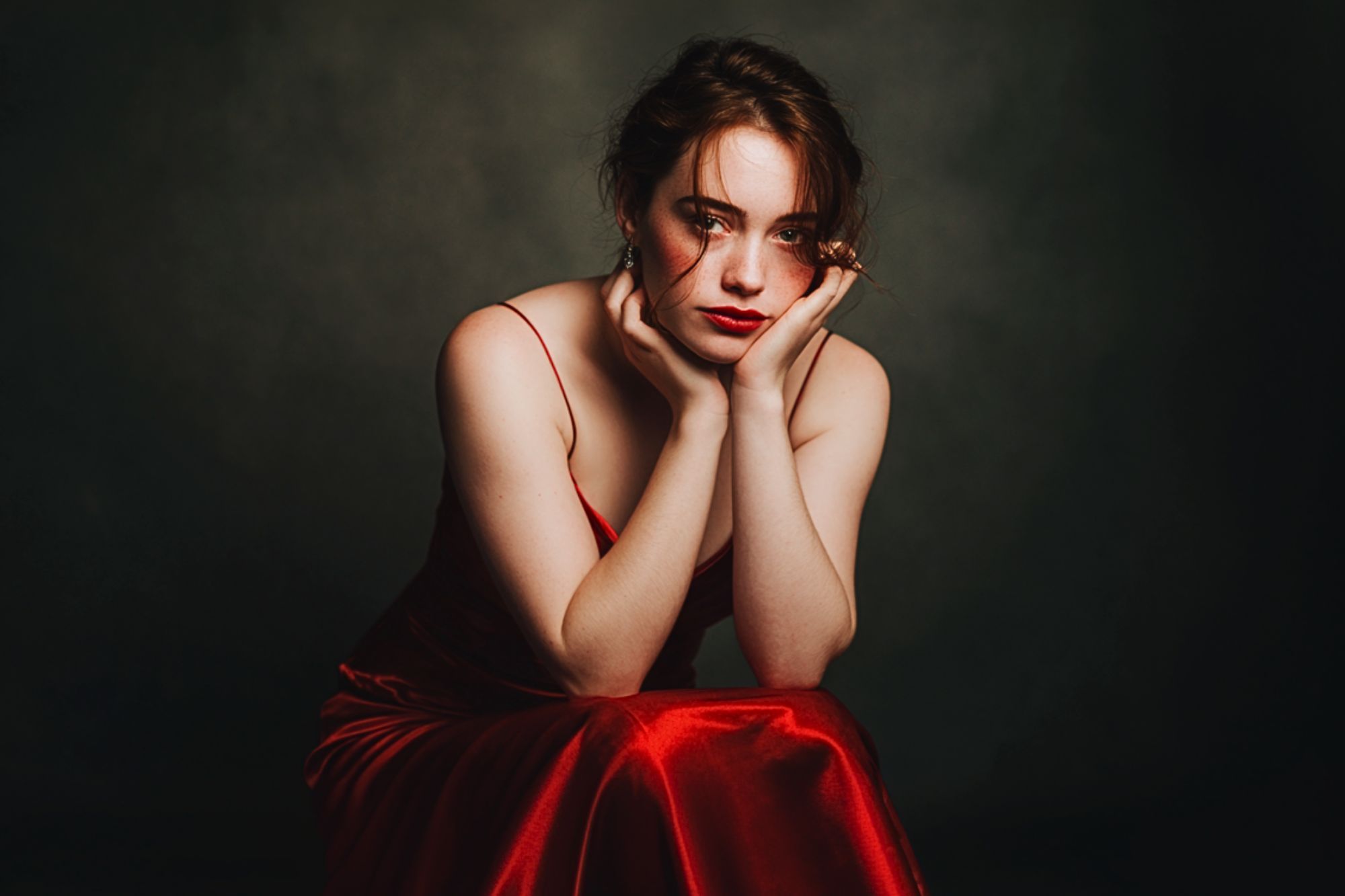 A woman in a red satin dress sits with her elbows on her knees, resting her chin on her hands, looking directly at the camera against a dark, blurred background.