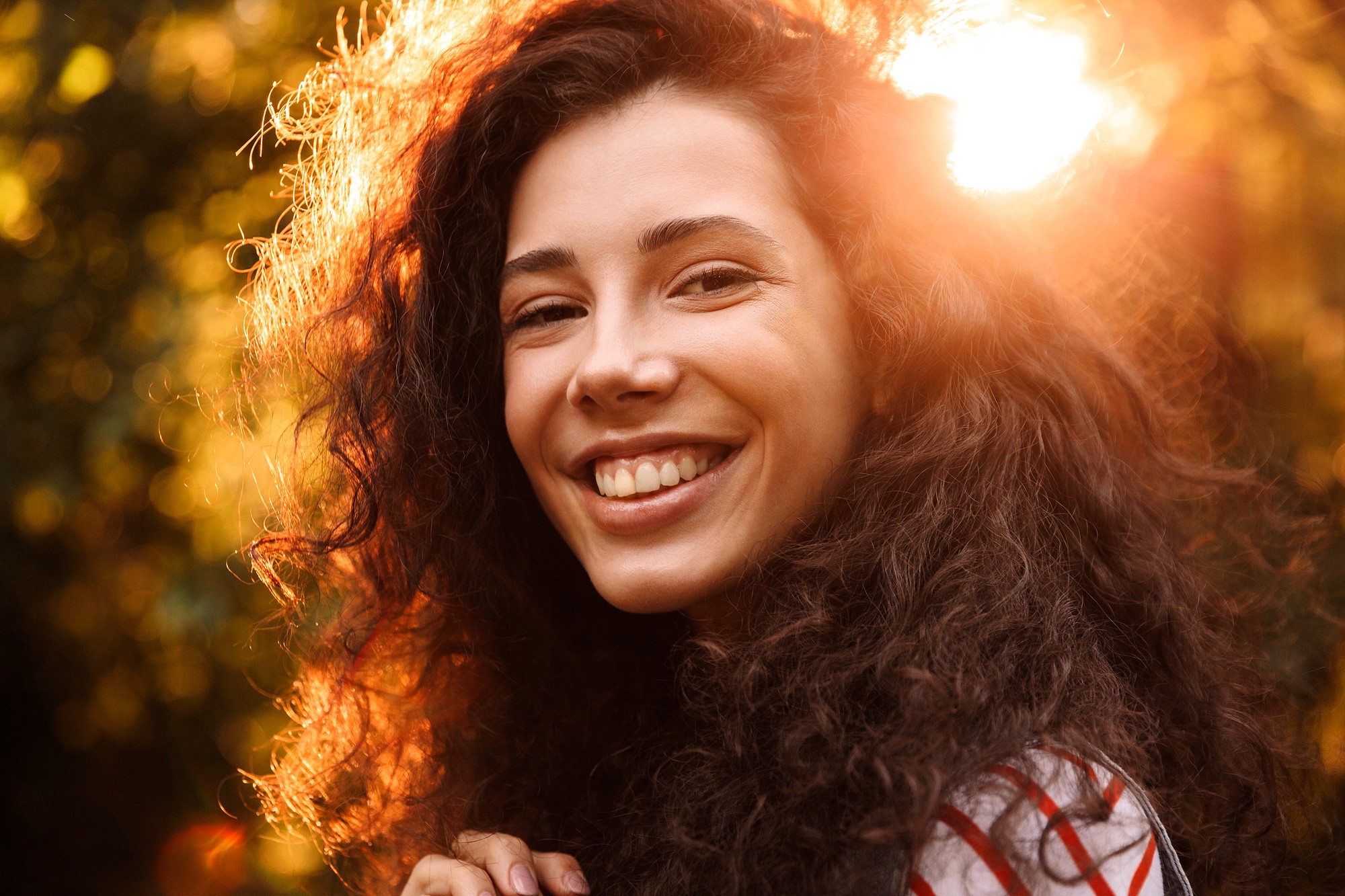 A woman with long, curly dark hair smiles warmly at the camera. She is outdoors with sunlight shining behind her, creating a golden, glowing effect around her head.