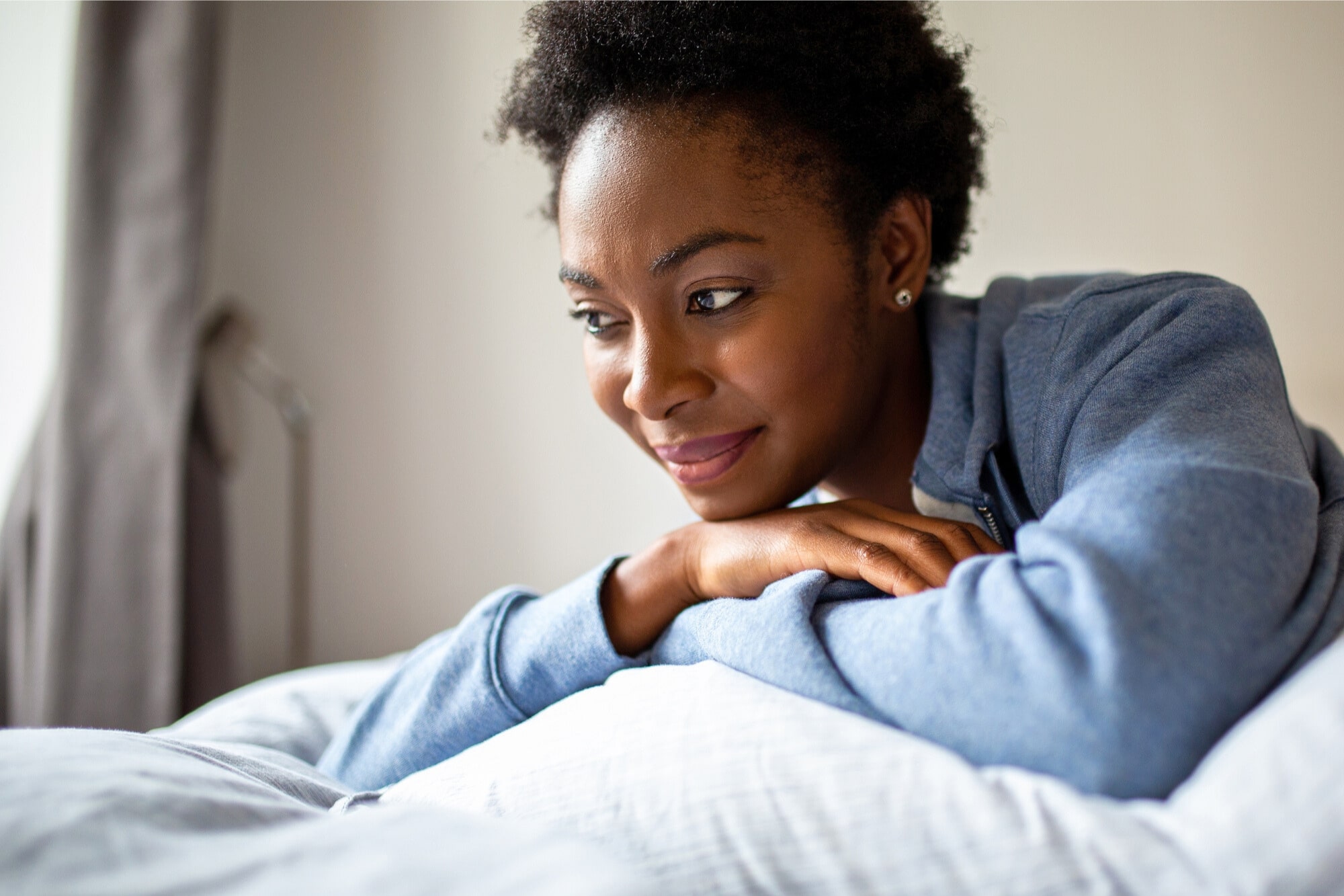 A woman in a blue hoodie rests her arms on a pillow, smiling softly and looking to the side in a bright, cozy room.