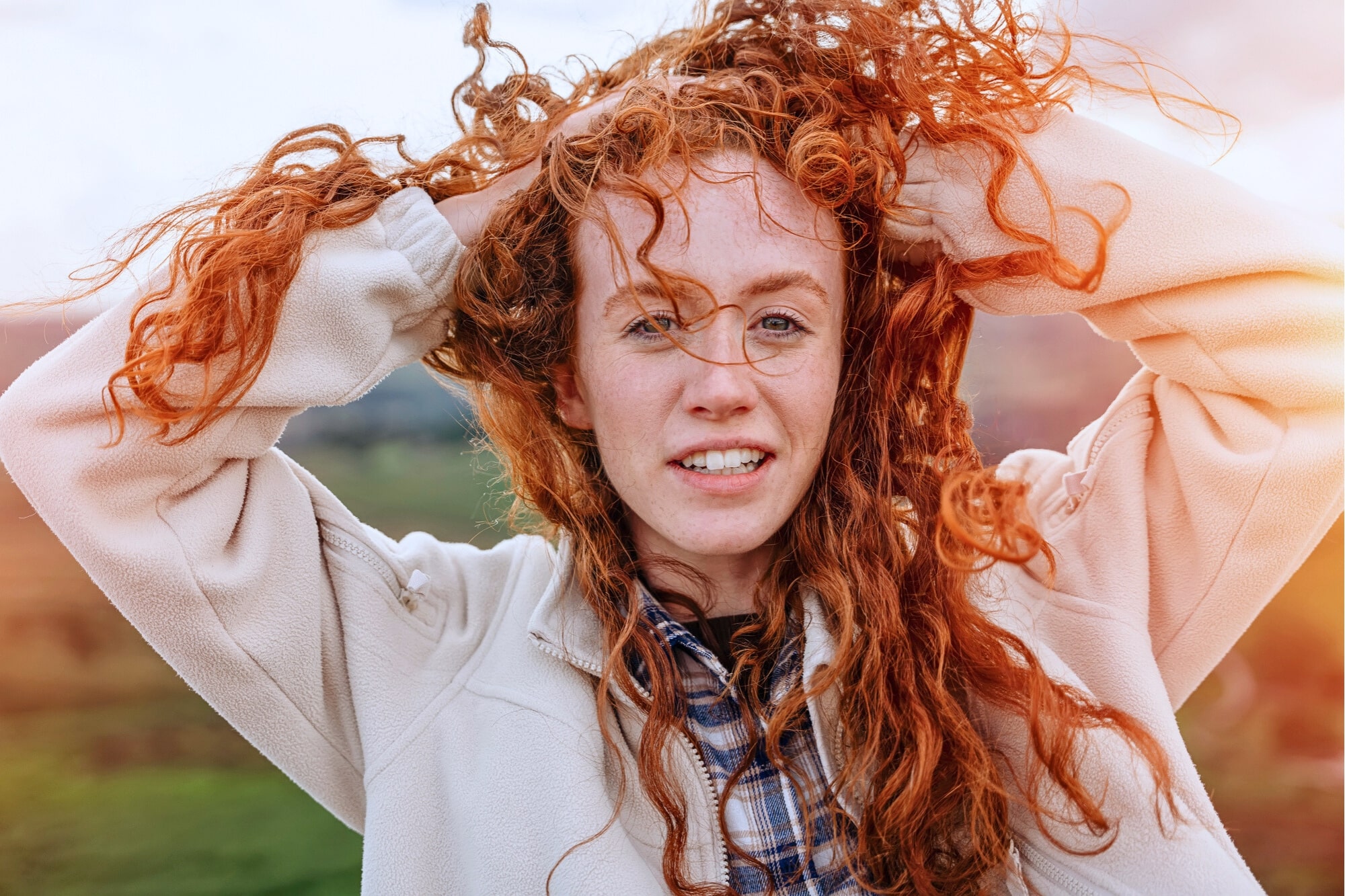 A young woman with long curly red hair holds her hands in her hair, smiling slightly. She wears a light jacket over a plaid shirt and stands outdoors with a natural, green landscape in the background.