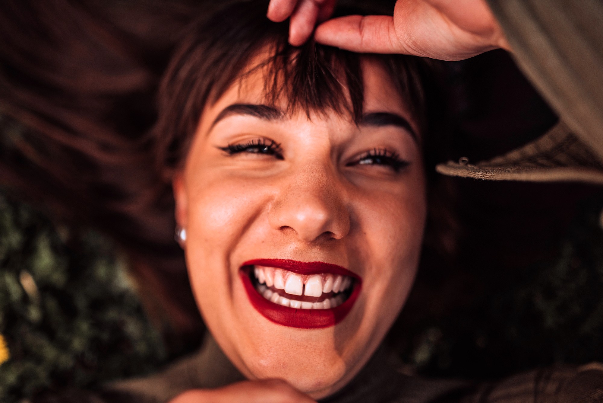 A person lying down, smiling widely with bright red lipstick and bold eyeliner, holding their bangs with one hand. The image captures a joyful, close-up expression.