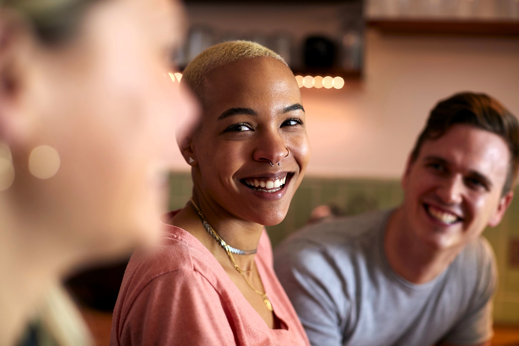 Three young adults are sitting together indoors, smiling and laughing. The focus is on a person with short blond hair and a nose ring, wearing a pink shirt, while the others are slightly out of focus in the foreground and background.