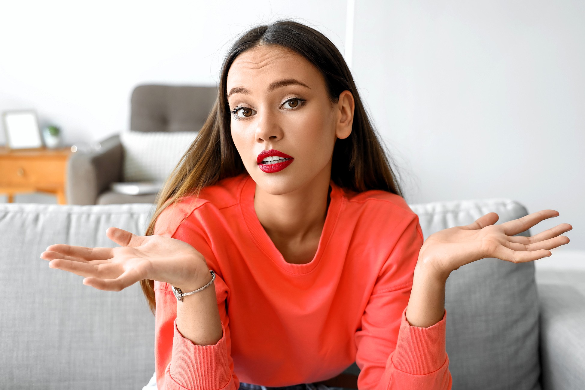 A young woman with long brown hair and red lipstick sits on a gray couch, wearing a coral sweatshirt, and raises both hands with an unsure or questioning expression. A blurred living room is in the background.