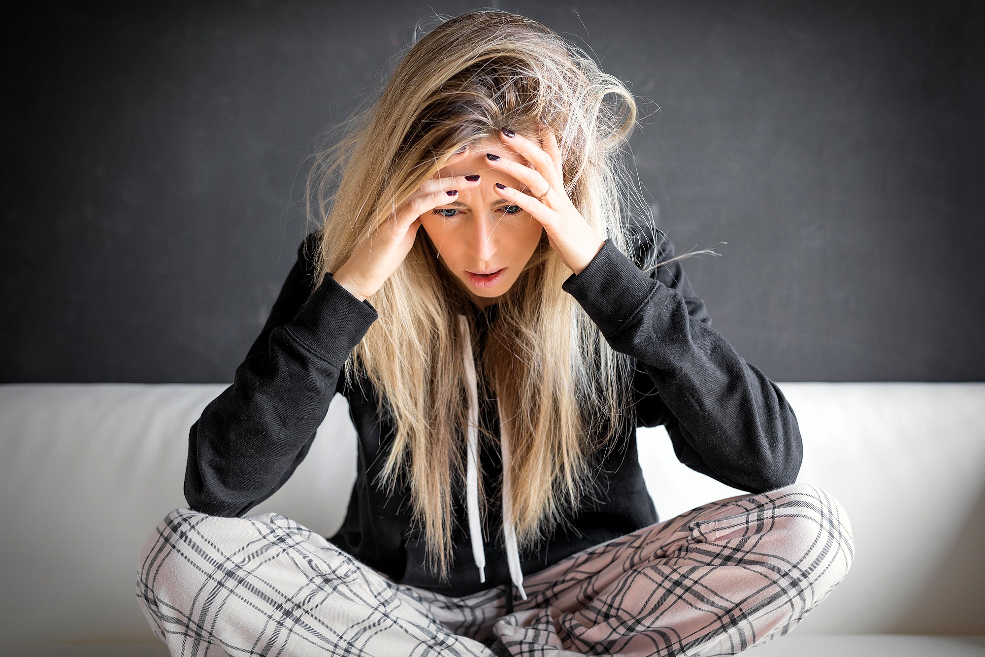 A young woman with long blonde hair sits cross-legged on a couch, wearing plaid pajama pants and a black hoodie, holding her head in her hands and looking down, appearing stressed or worried.