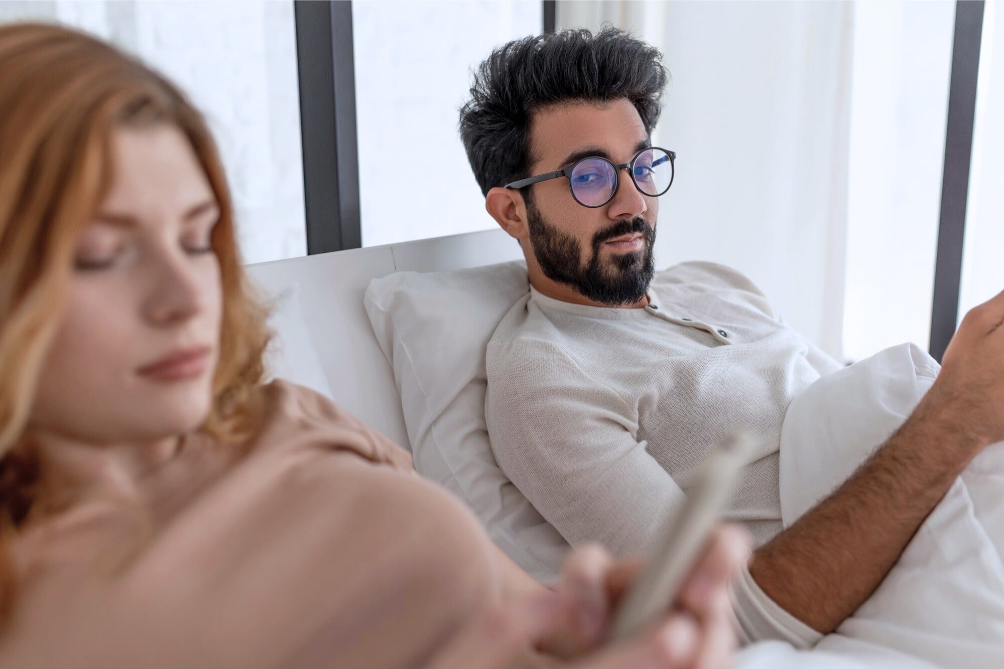 A man and woman lie in bed using their phones. The man, wearing glasses, looks over at the woman, who is focused on her phone. Both appear relaxed. The background is bright with natural light.