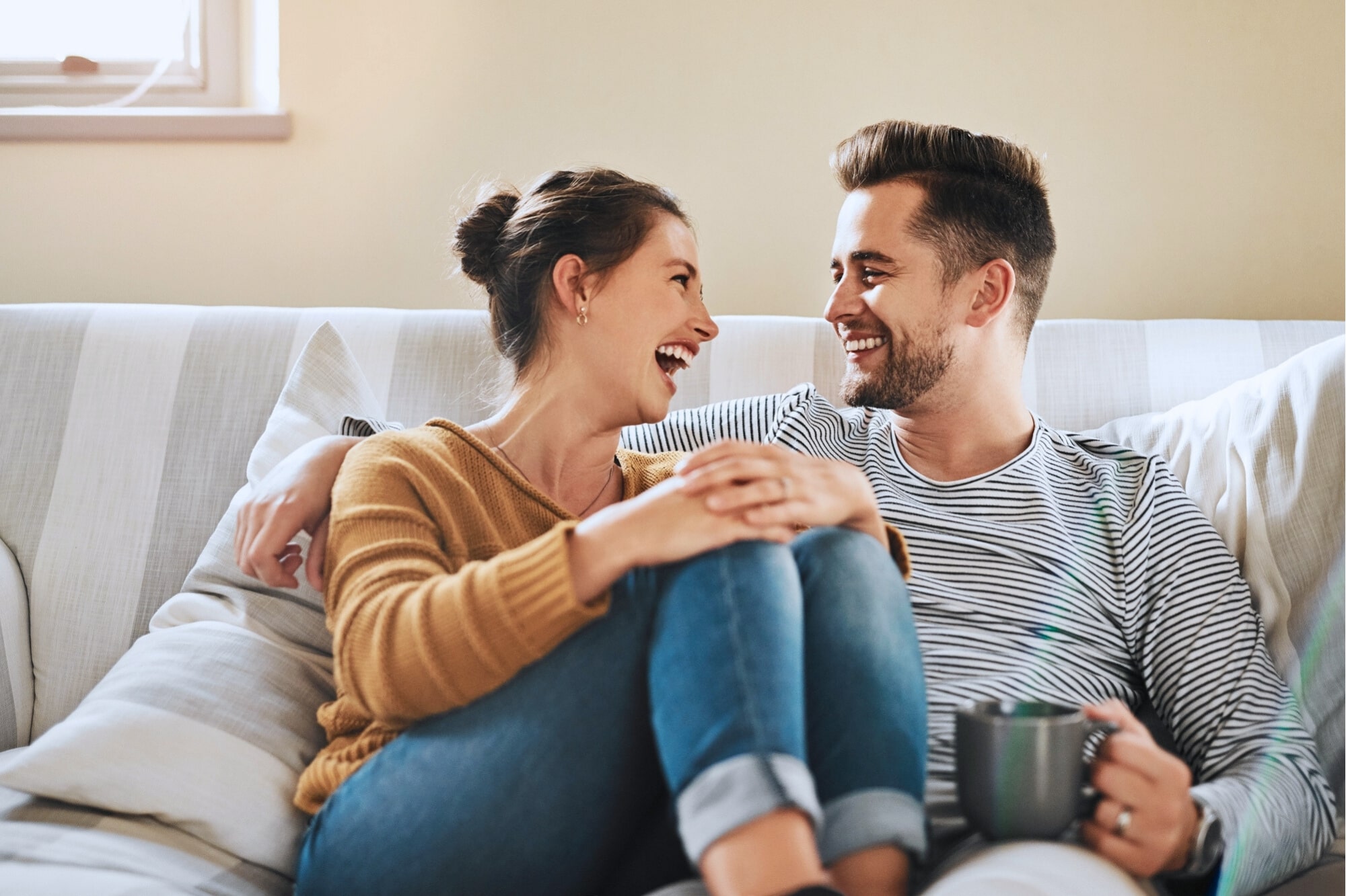 A smiling couple sits close together on a couch, laughing and looking at each other. One person wears a mustard sweater and jeans, while the other wears a striped shirt and holds a mug. The atmosphere appears warm and relaxed.