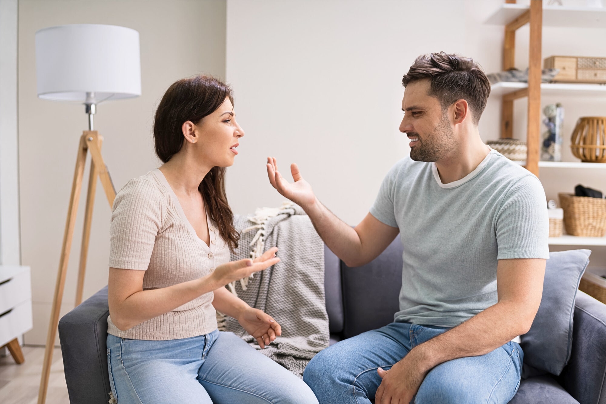 A woman and man sit on a couch in a living room, facing each other and appearing to have an intense discussion or argument, with expressive hand gestures and concerned facial expressions.