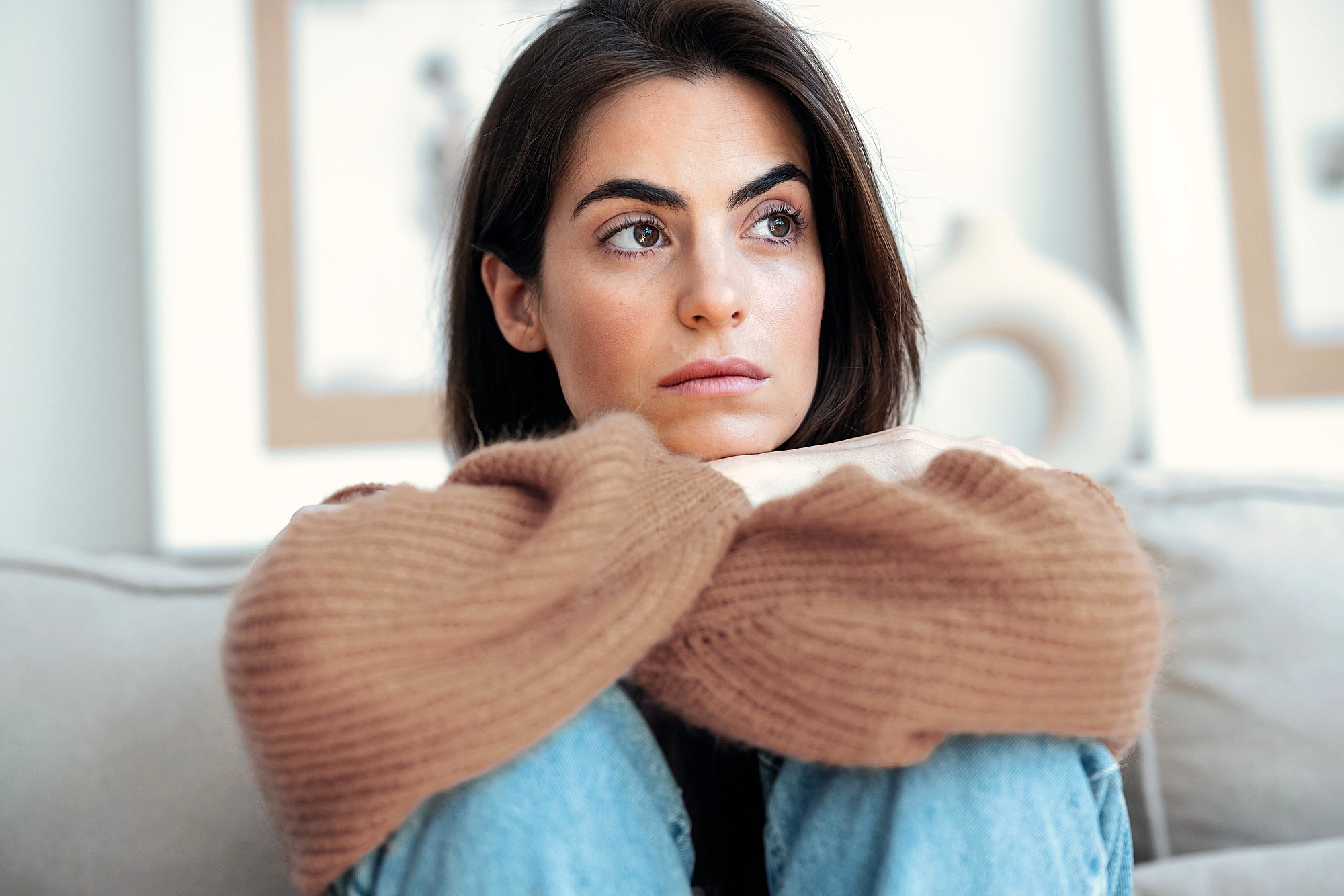 A woman with long dark hair sits on a couch, resting her chin on her crossed arms. She wears a brown sweater and blue jeans, and looks thoughtfully to the side. The background is softly blurred.