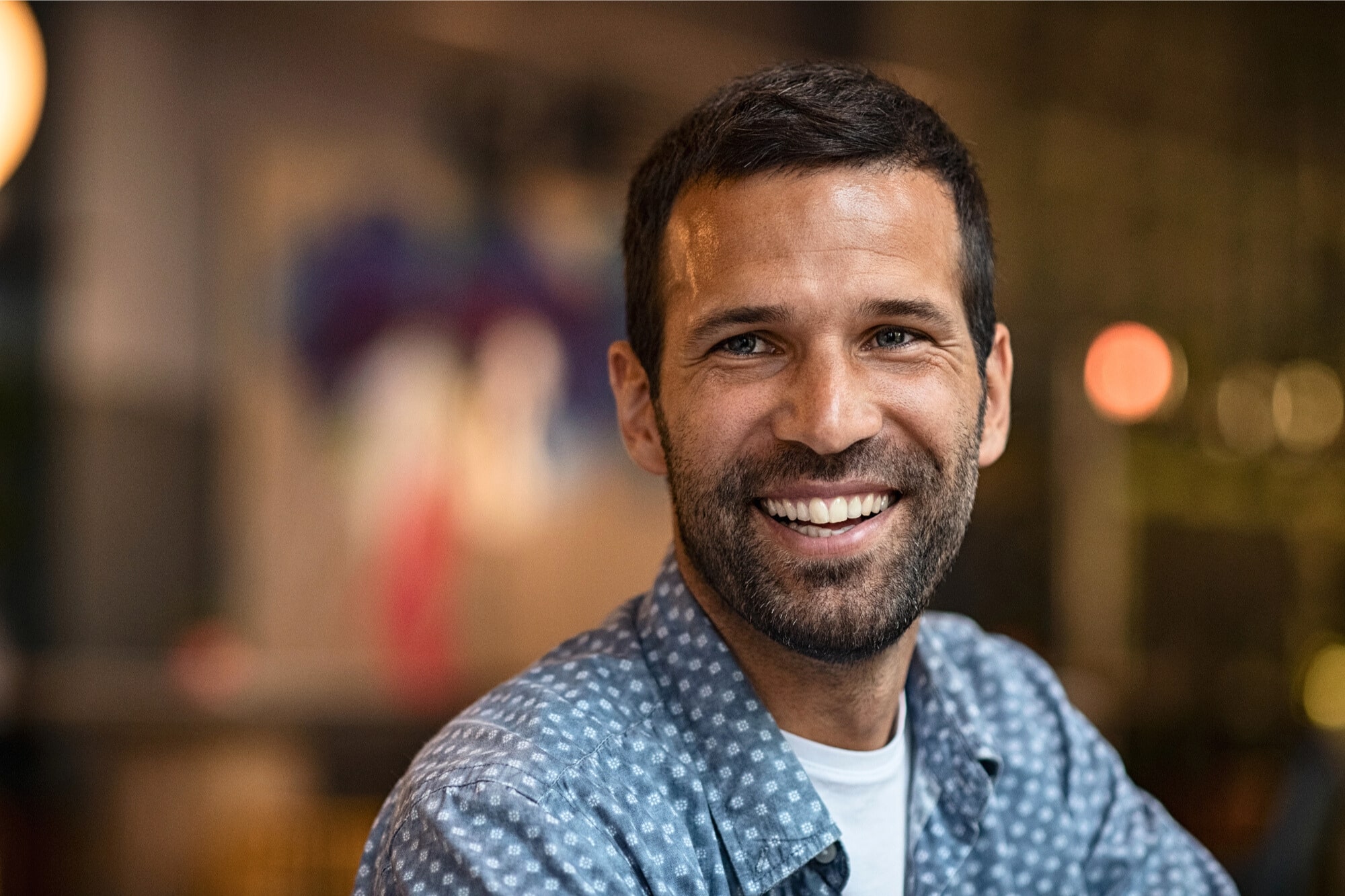 A man with short dark hair and a beard smiles warmly at the camera, wearing a blue patterned shirt. The background is softly blurred, suggesting an indoor setting with ambient lighting.