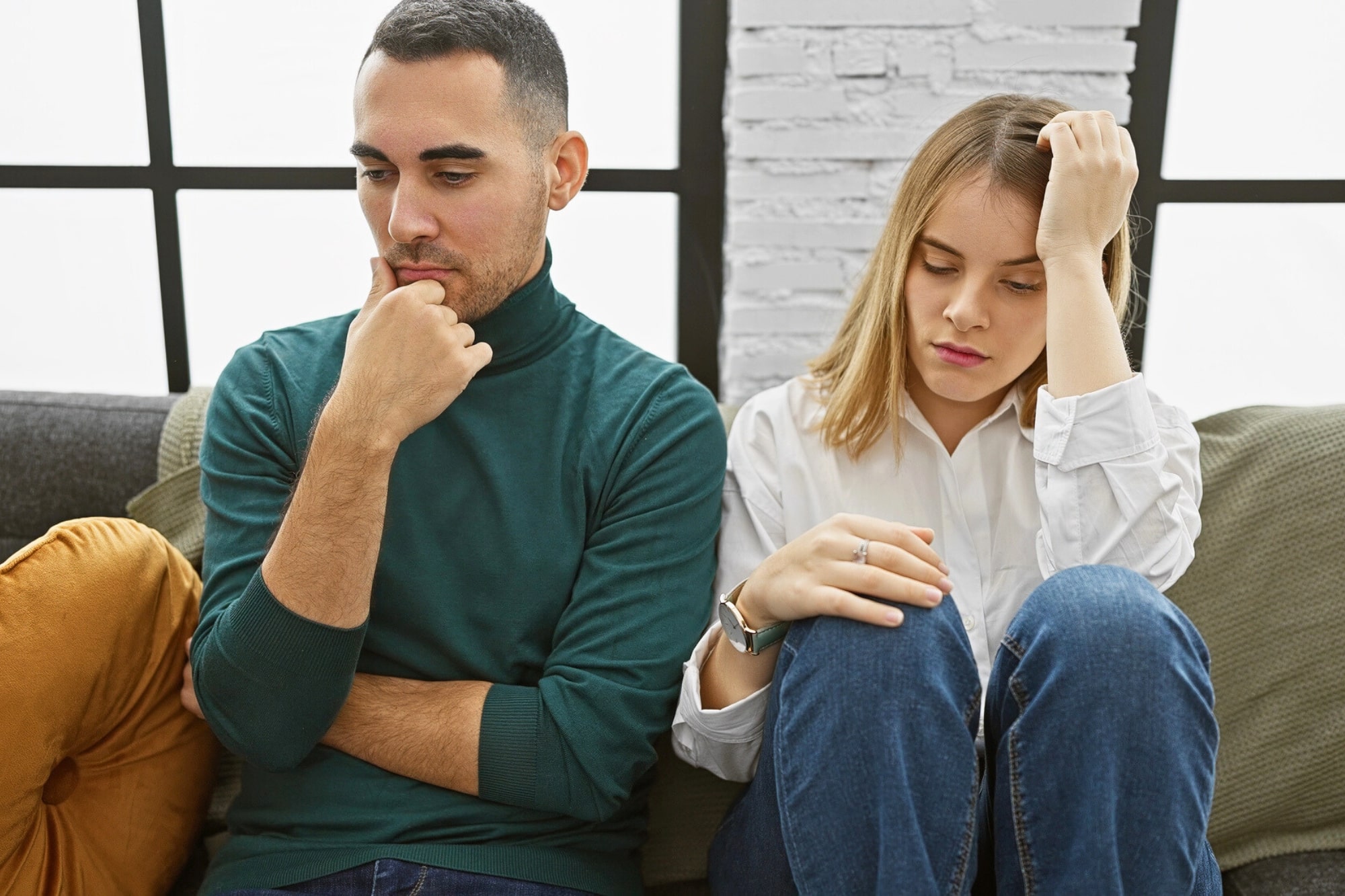 A man and a woman sit on a couch looking upset and distant. The man rests his chin on his hand, while the woman holds her forehead and looks down. Both appear to be deep in thought or troubled.
