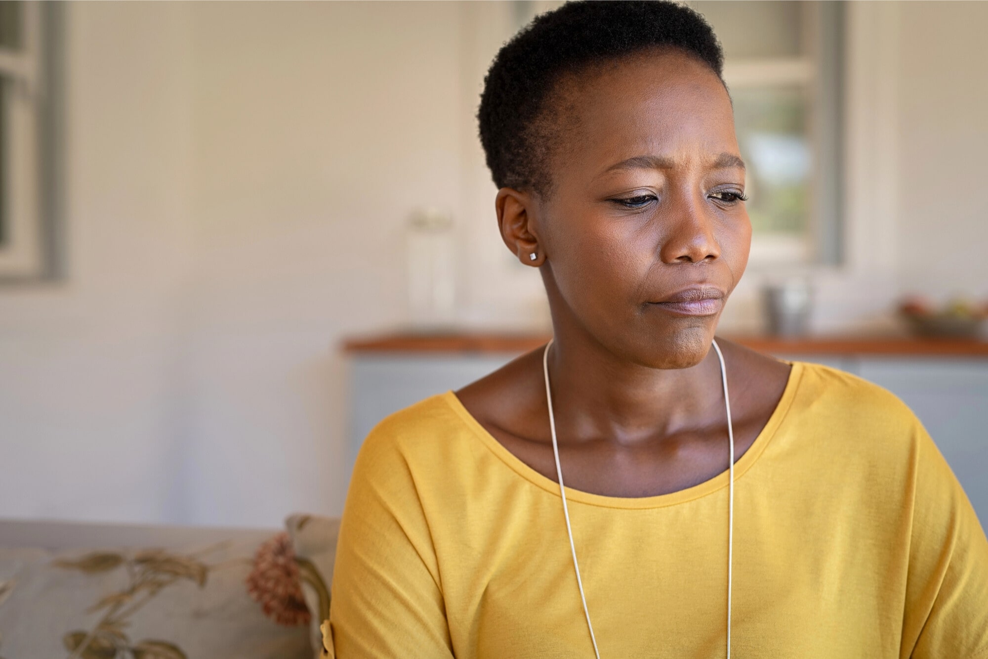 A woman wearing a yellow top sits indoors, looking down with a thoughtful or concerned expression. The background is softly lit and out of focus.