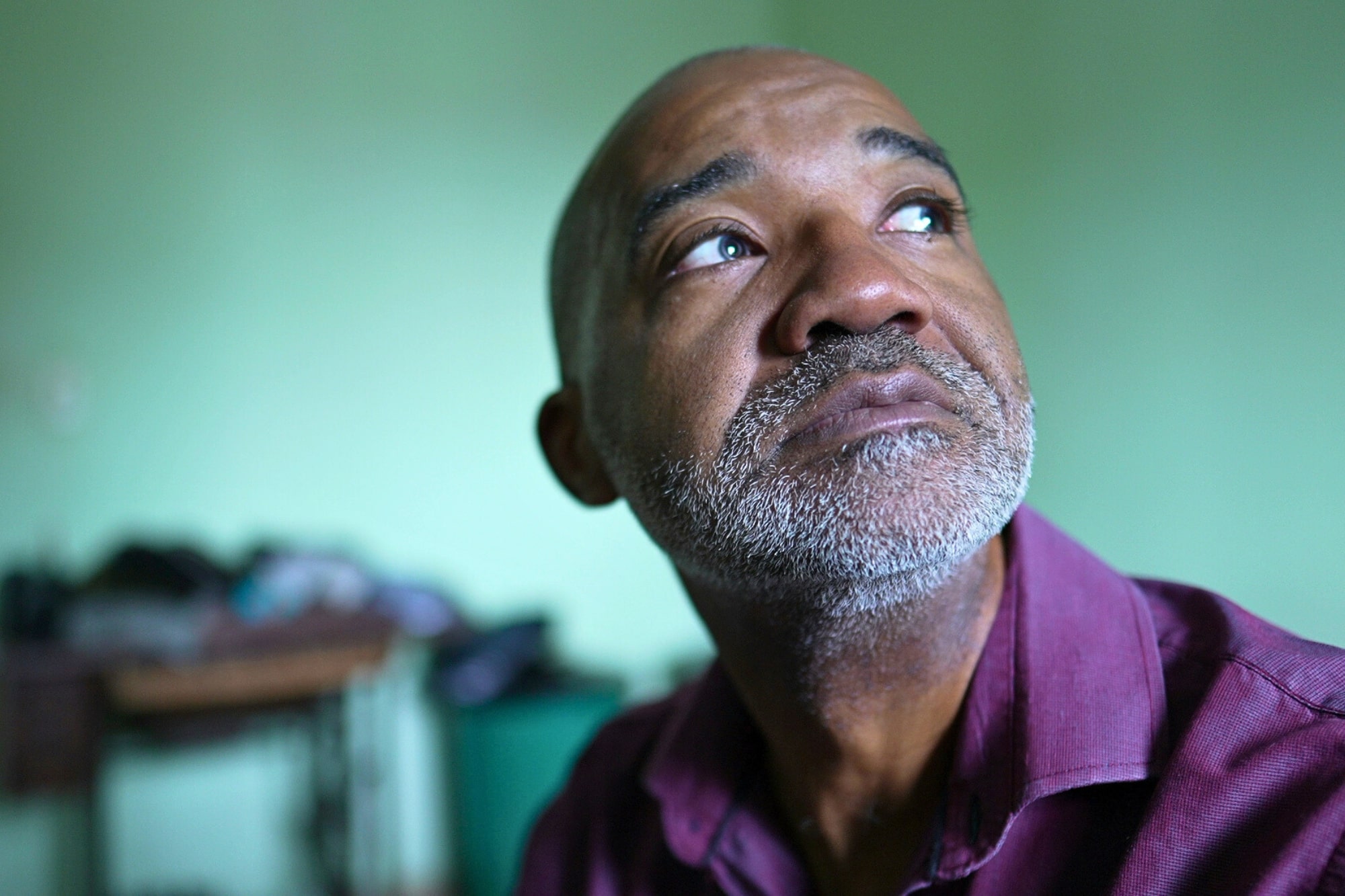 A middle-aged man with a graying beard and mustache looks thoughtfully upward. He is wearing a maroon shirt and is indoors, with a soft green background and some blurred furniture behind him.