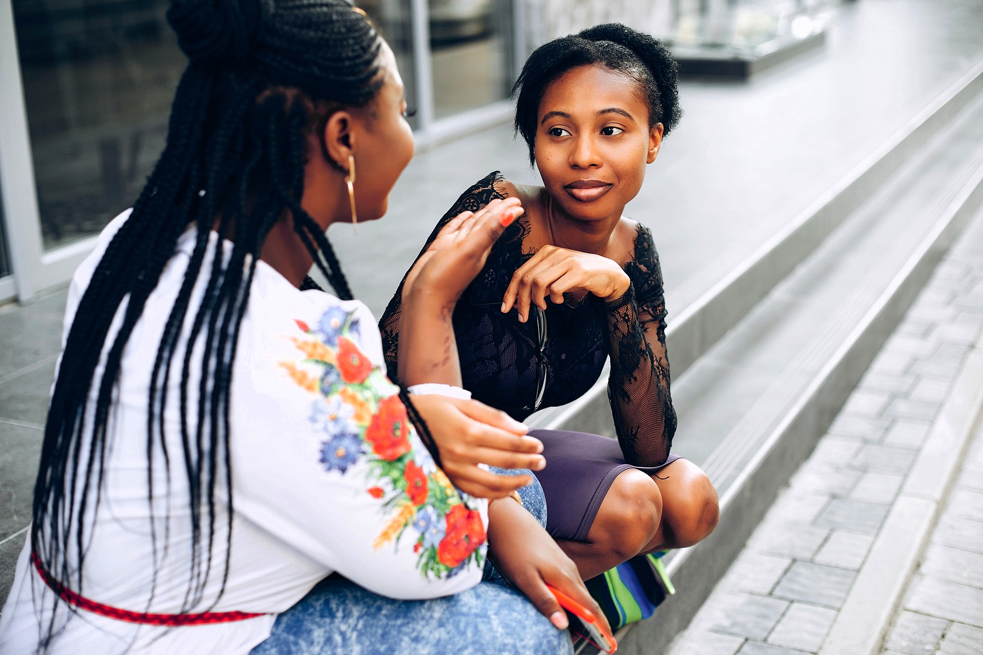Two women sit on outdoor steps, engaged in a lively conversation. One woman gestures with her hand while the other listens attentively, smiling. Both are casually dressed, and the scene is bright and relaxed.