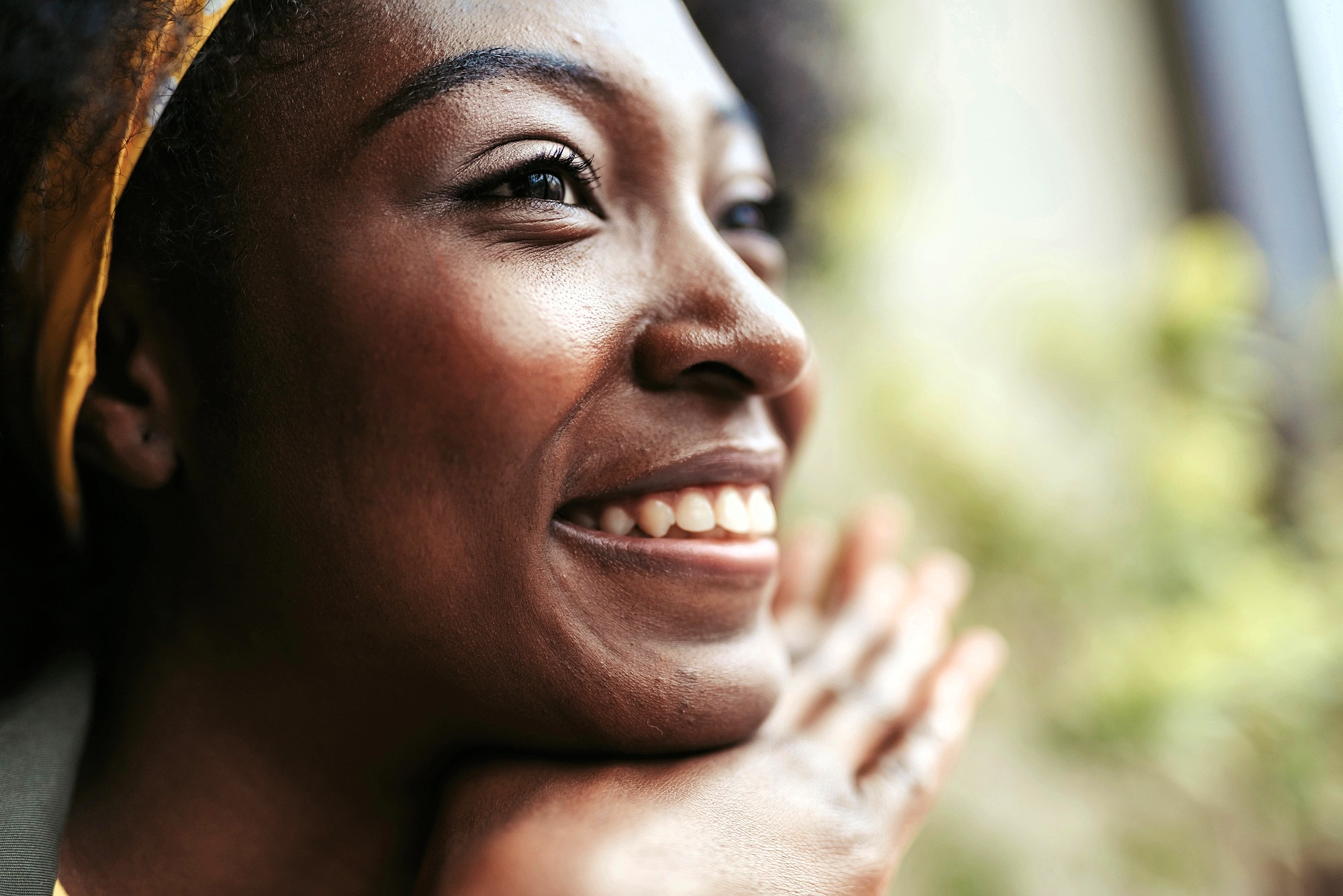 A close-up of a smiling woman resting her chin on her hands, looking off to the side with a joyful expression. Bright natural light highlights her face, and a blurred, green background is visible.