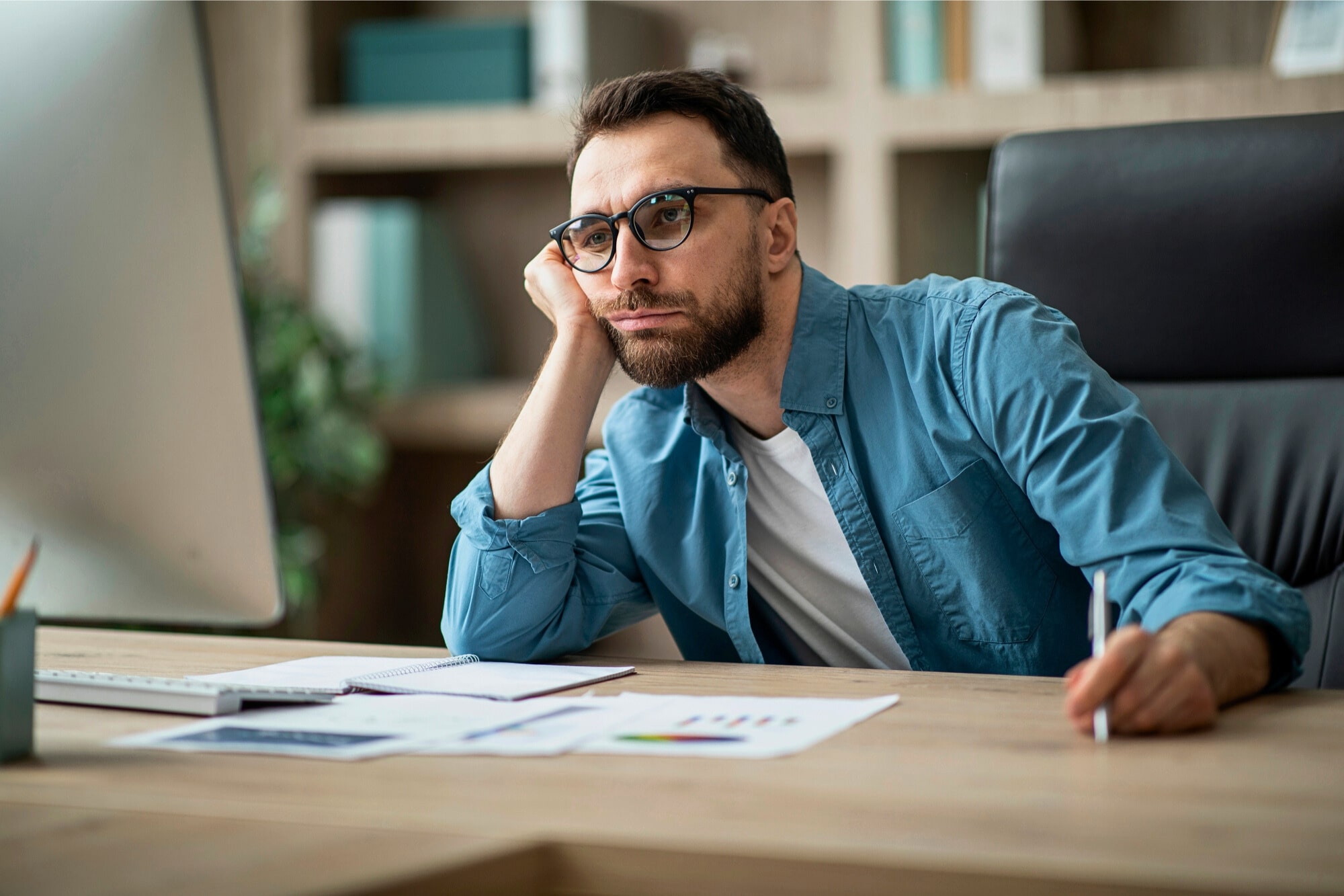 A man with glasses and a beard sits at a desk, resting his chin on his hand and looking thoughtfully at a computer screen. Papers, a pen, and office supplies are spread out in front of him.