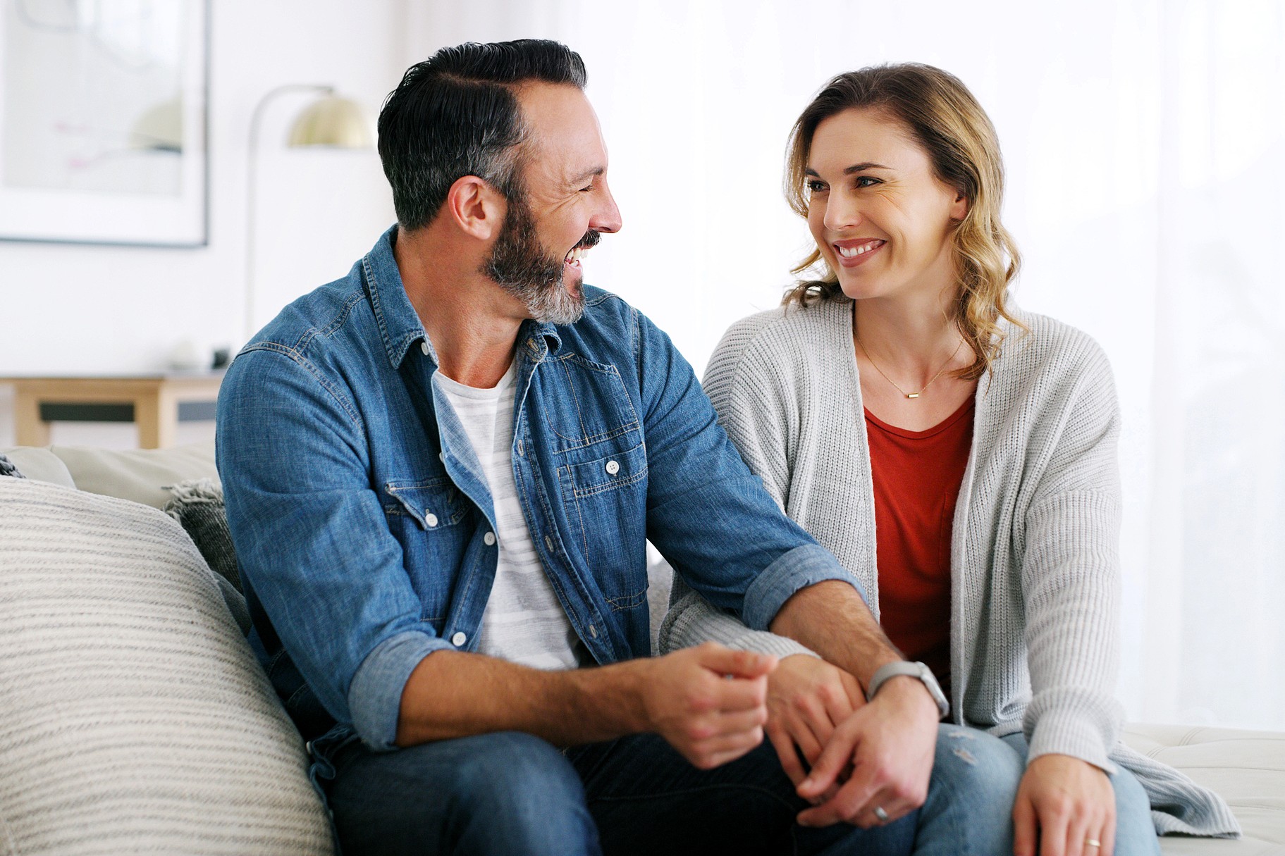 A smiling man and woman sit closely together on a couch, holding hands and looking at each other warmly in a cozy, well-lit living room.