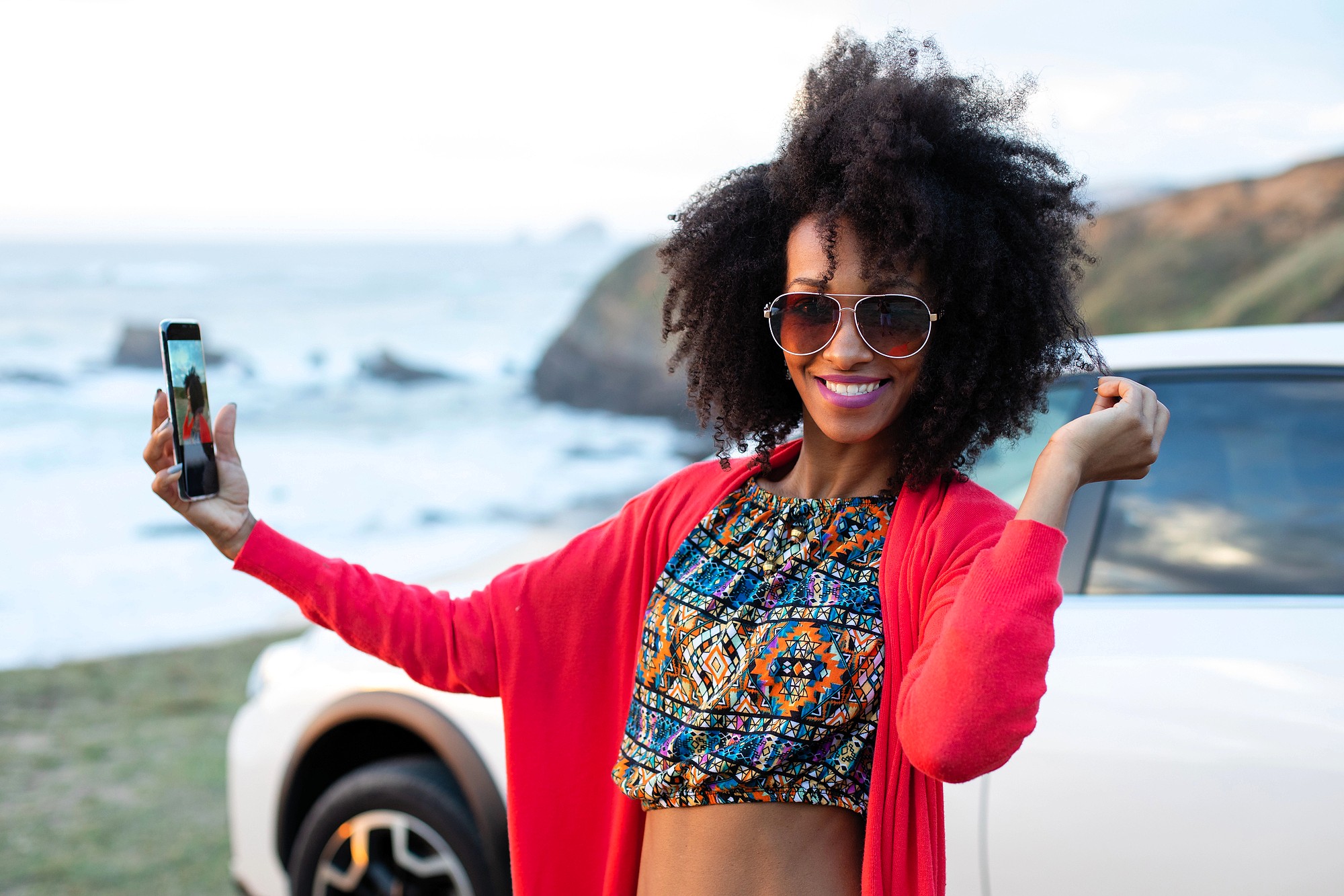 A smiling woman wearing sunglasses and a colorful crop top takes a selfie by the seaside, standing next to a white car with cliffs and ocean in the background.