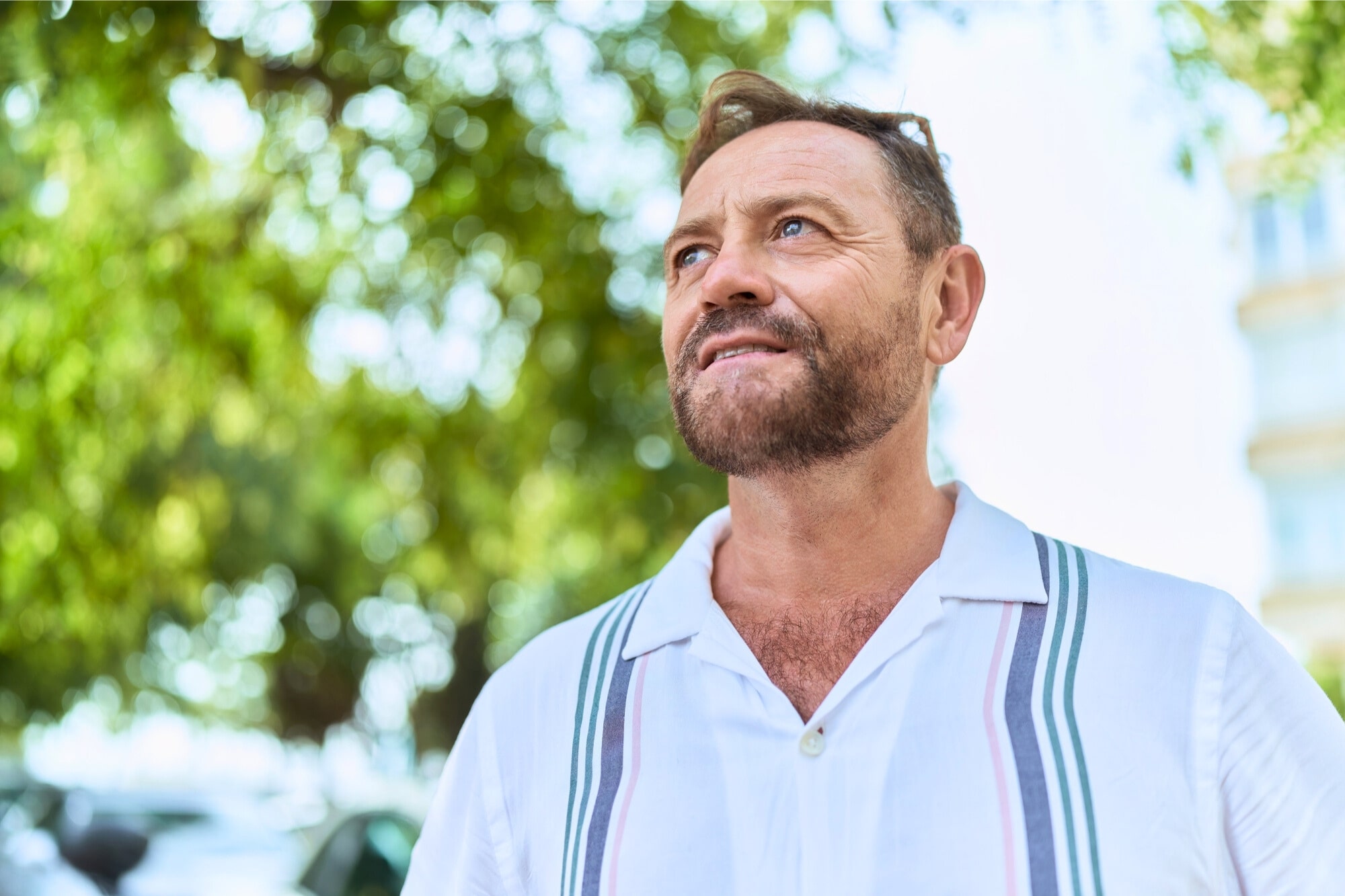 A middle-aged man with a beard and short hair, wearing a white shirt with stripes, stands outdoors and looks thoughtfully into the distance. Blurred green trees and sunlight are in the background.
