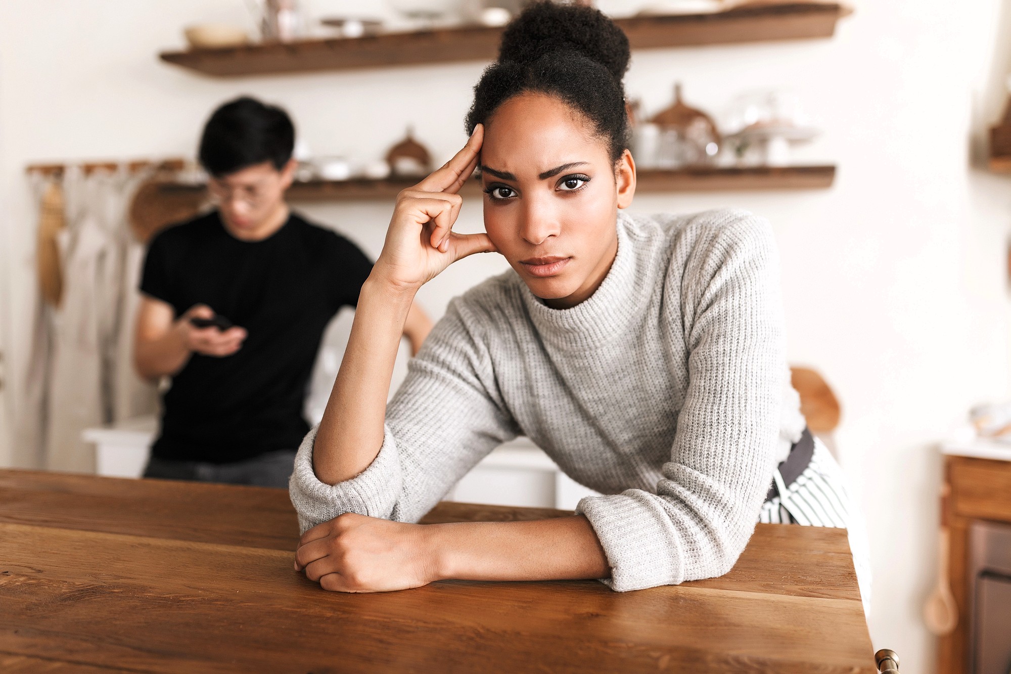 A woman in a gray sweater leans on a wooden table, looking serious and thoughtful. In the background, a man stands using his phone, slightly out of focus. The setting appears to be a cozy kitchen.