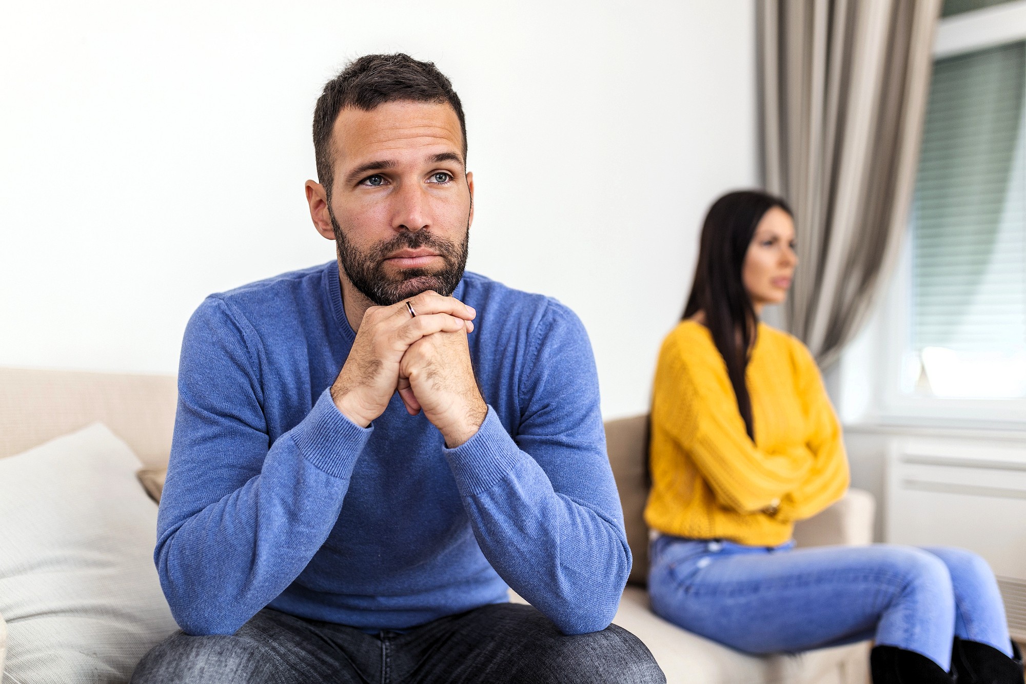 A man in a blue sweater sits on a couch, looking thoughtful with his hands clasped under his chin. Behind him, a woman in a yellow sweater sits with her arms crossed, facing away. Both appear upset.