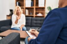 A person sits on a couch in conversation with another person holding a clipboard and pen, suggesting a therapy or counseling session in a comfortable office setting.