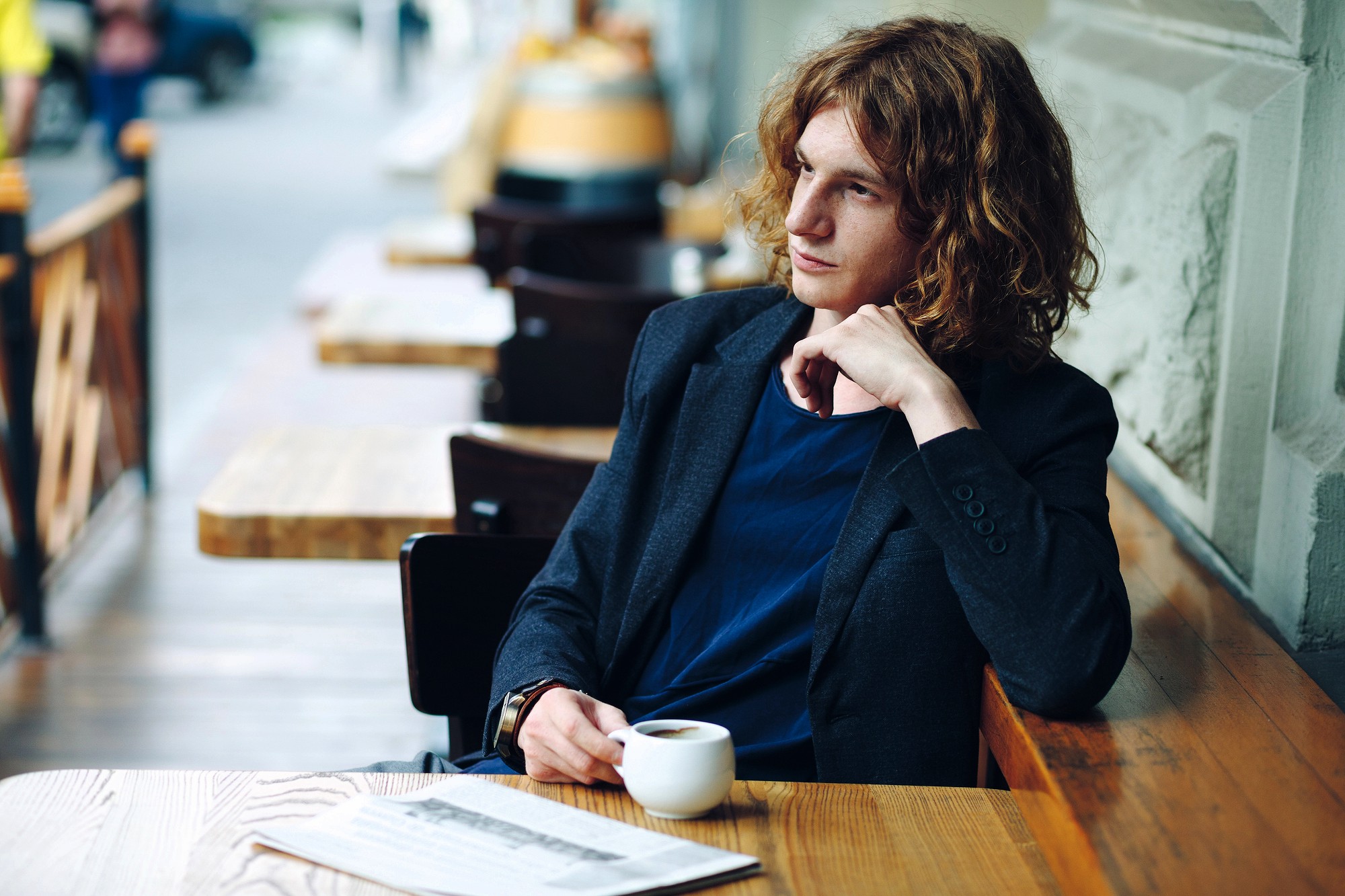 A person with wavy, shoulder-length hair sits at an outdoor café table, holding a cup of coffee and gazing thoughtfully to the side. A newspaper is on the table in front of them.