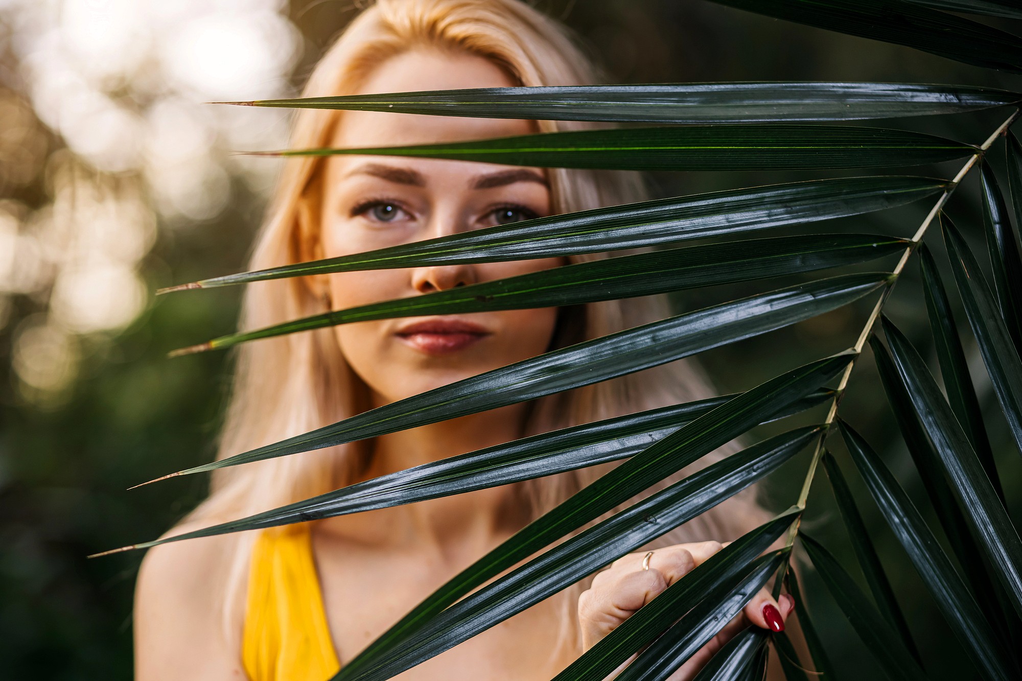 A woman with long blonde hair stands behind green palm leaves, with her face partially obscured. She is wearing a yellow top and has red painted nails. The background is softly blurred with natural light.