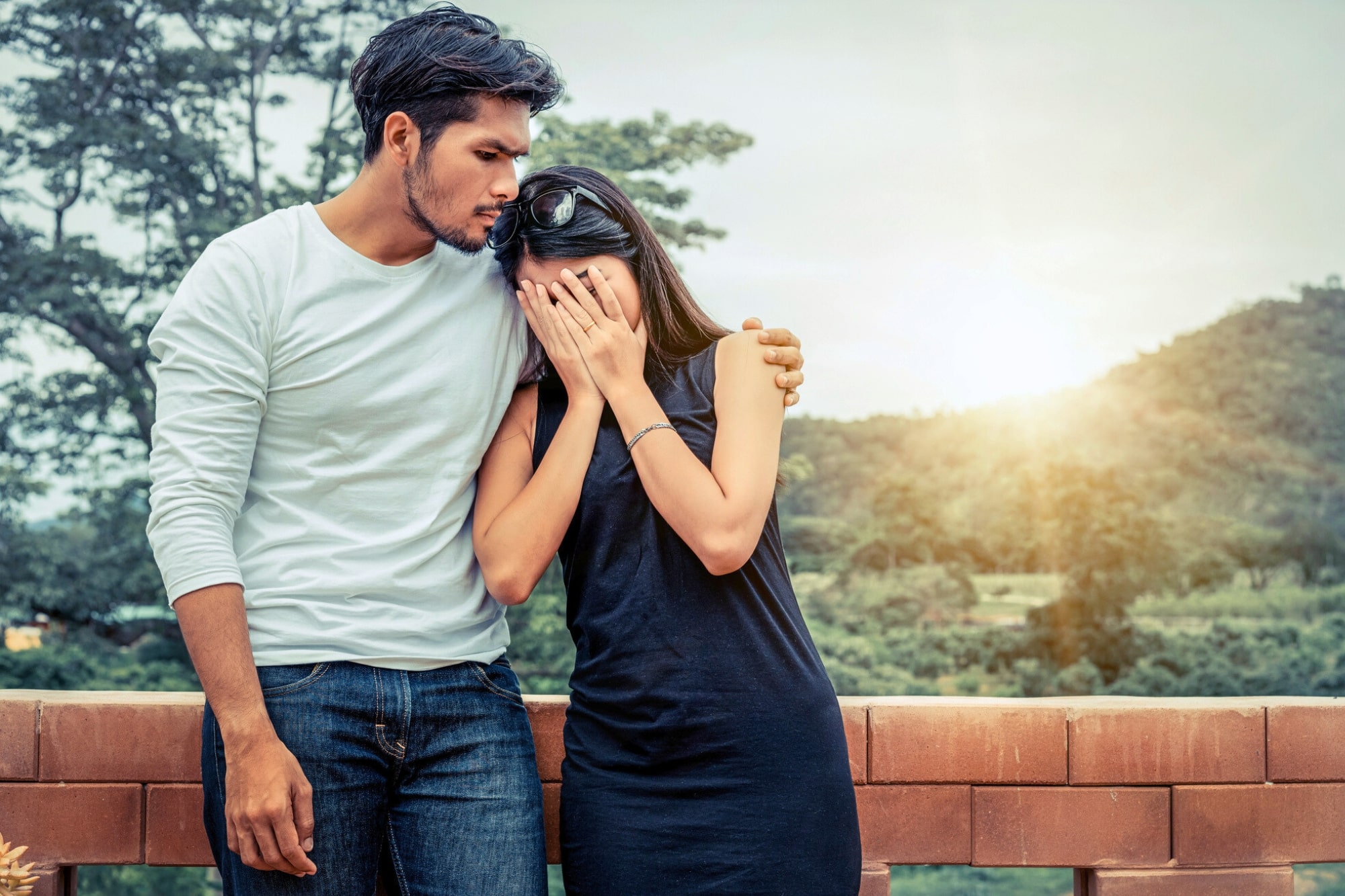 A man in a white shirt comforts a woman in a black dress who is covering her face with her hands. They are standing outdoors with trees and hills in the background at sunset.