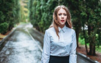 A young woman with long, light brown hair stands on a wet, tree-lined road, wearing a white button-up shirt and black skirt, looking directly at the camera with a neutral expression.