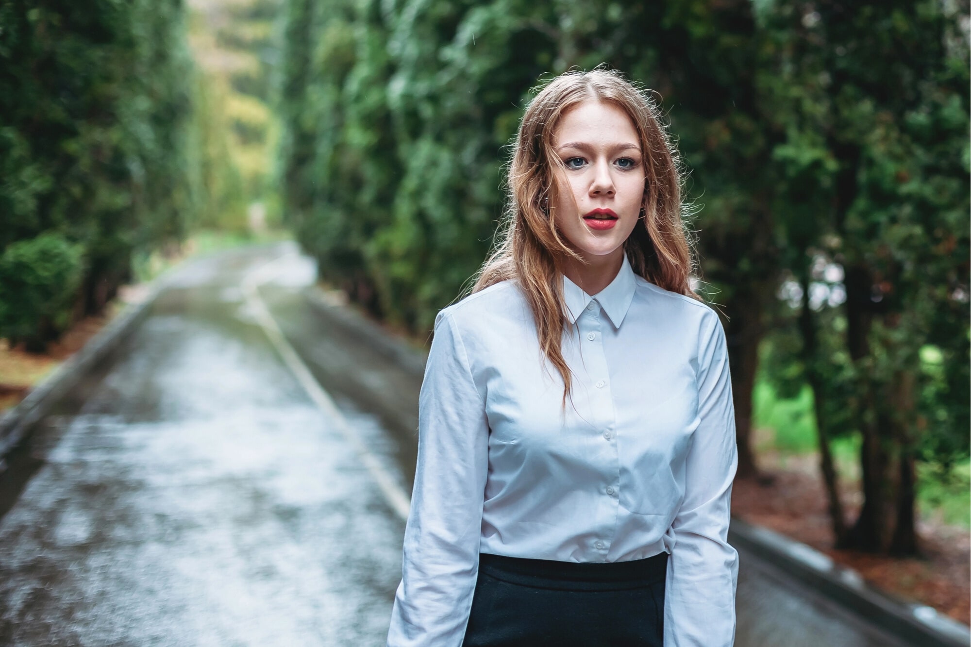 A young woman with long, light brown hair stands on a wet, tree-lined road, wearing a white button-up shirt and black skirt, looking directly at the camera with a neutral expression.