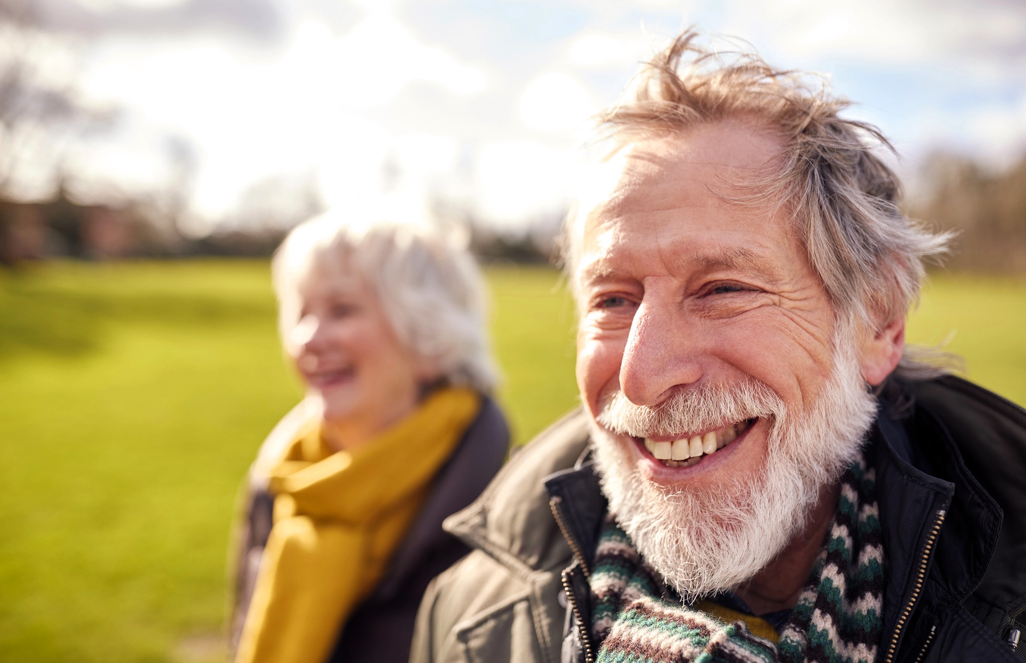 An older man with a gray beard smiles outdoors, wearing a striped scarf and jacket. A woman with gray hair and a yellow scarf stands slightly behind him. Both appear happy in a sunlit, grassy park.