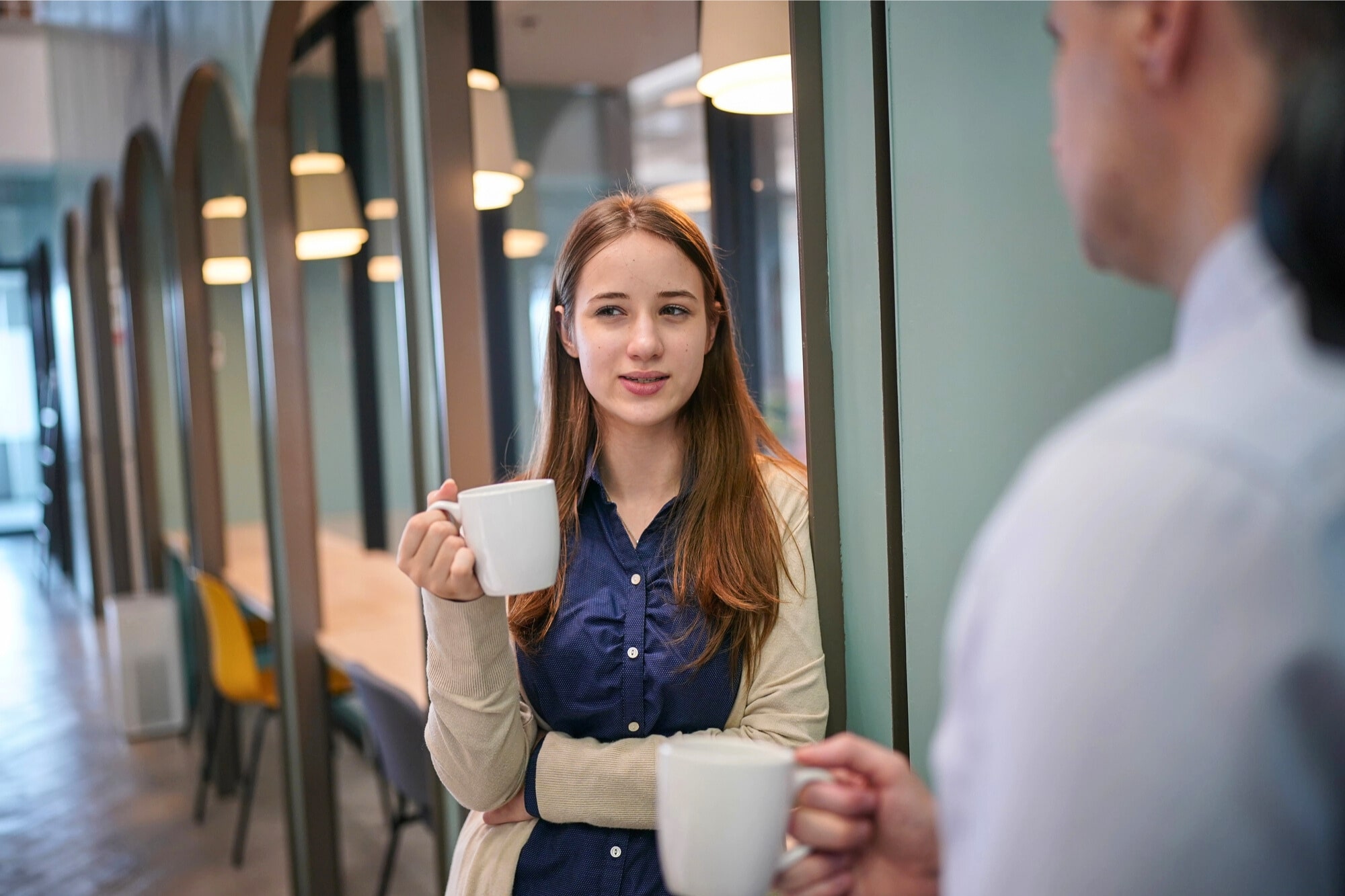 A young woman holding a white mug stands in a modern office hallway, talking to a person off to the right who is also holding a mug. She looks engaged in the conversation.