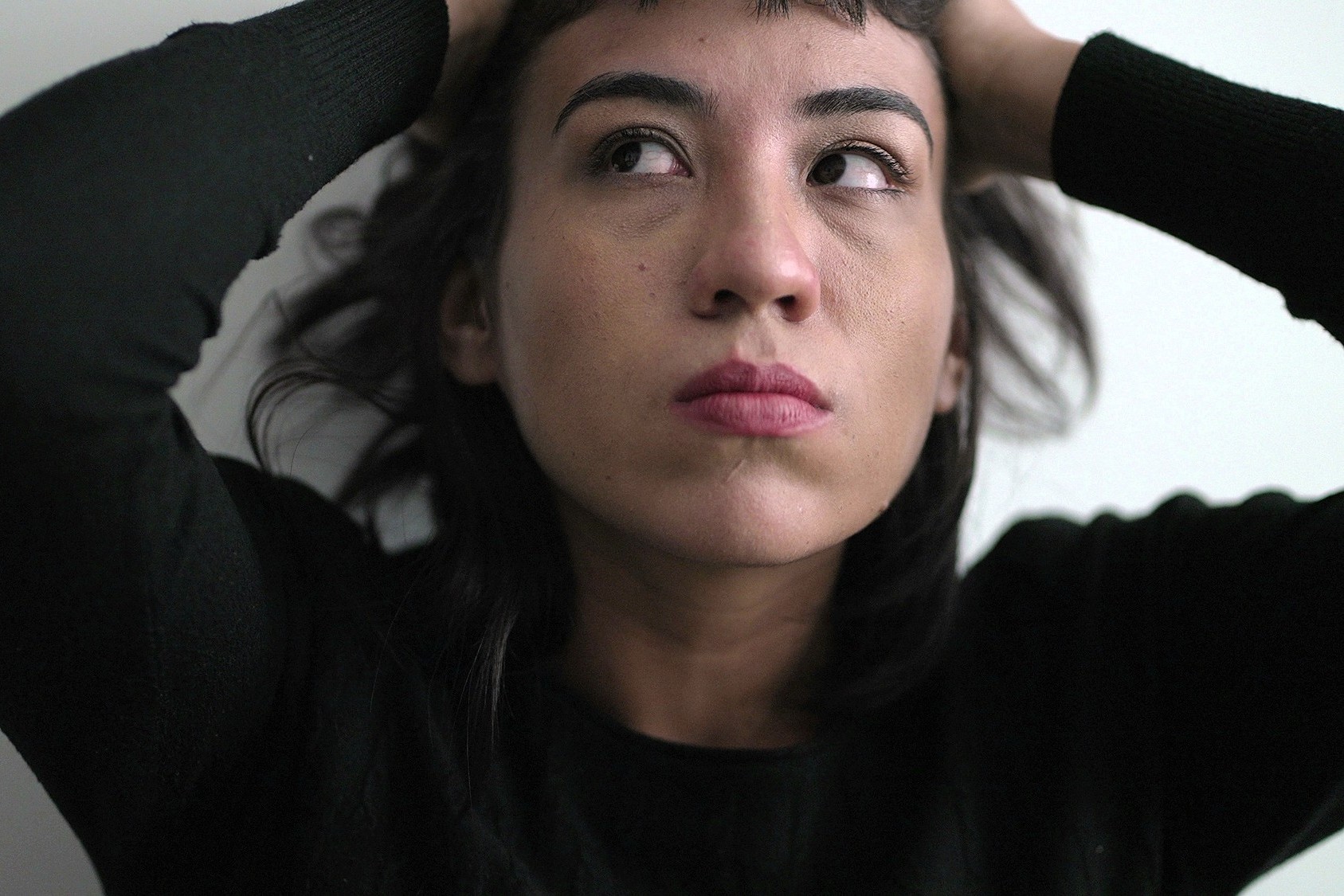 A person with dark hair and a serious expression looks upward, holding their hands on their head. They are wearing a black top, and the background is plain and light-colored.